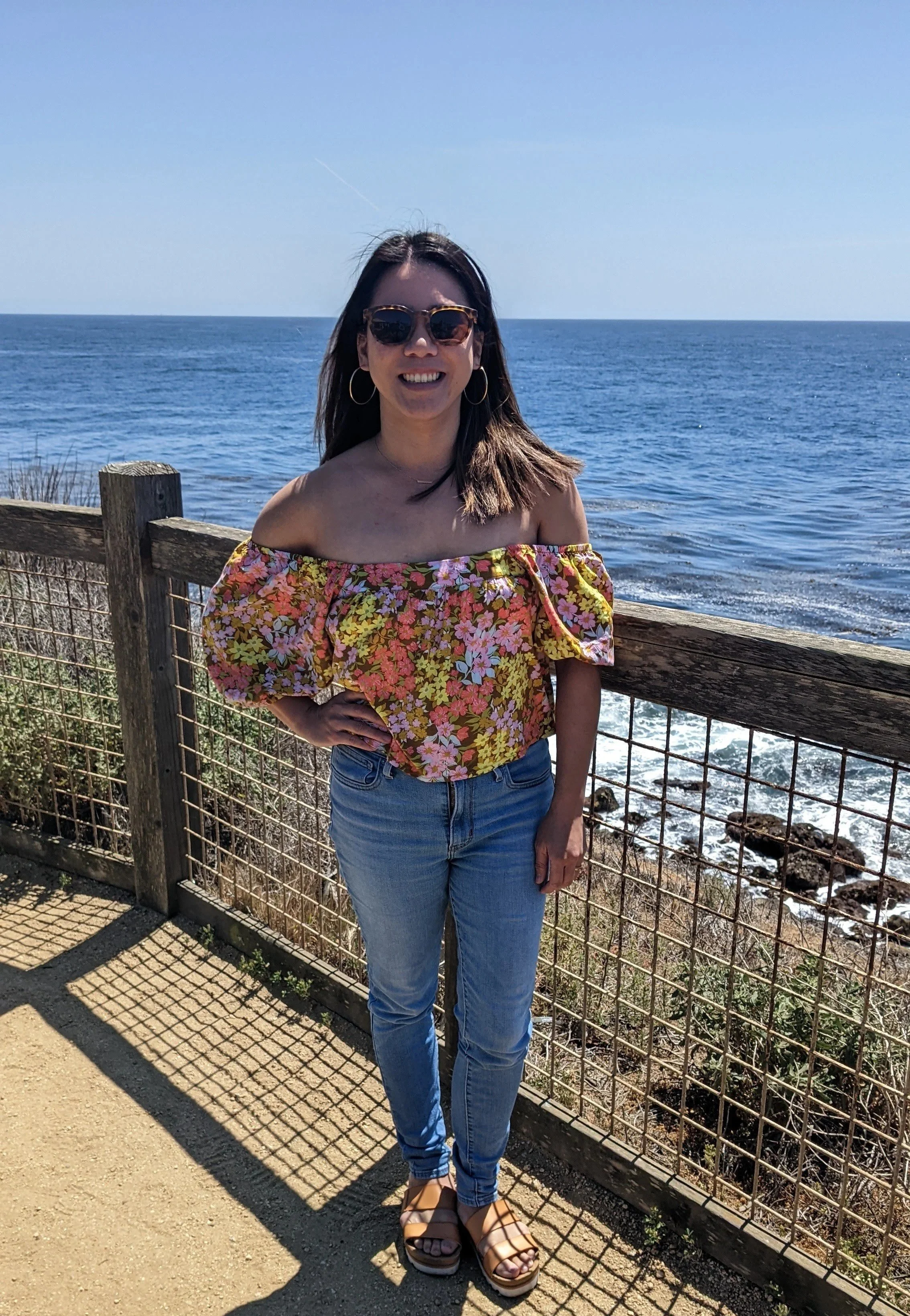 Woman in floral off-shoulder top and jeans standing by ocean railing on sunny day