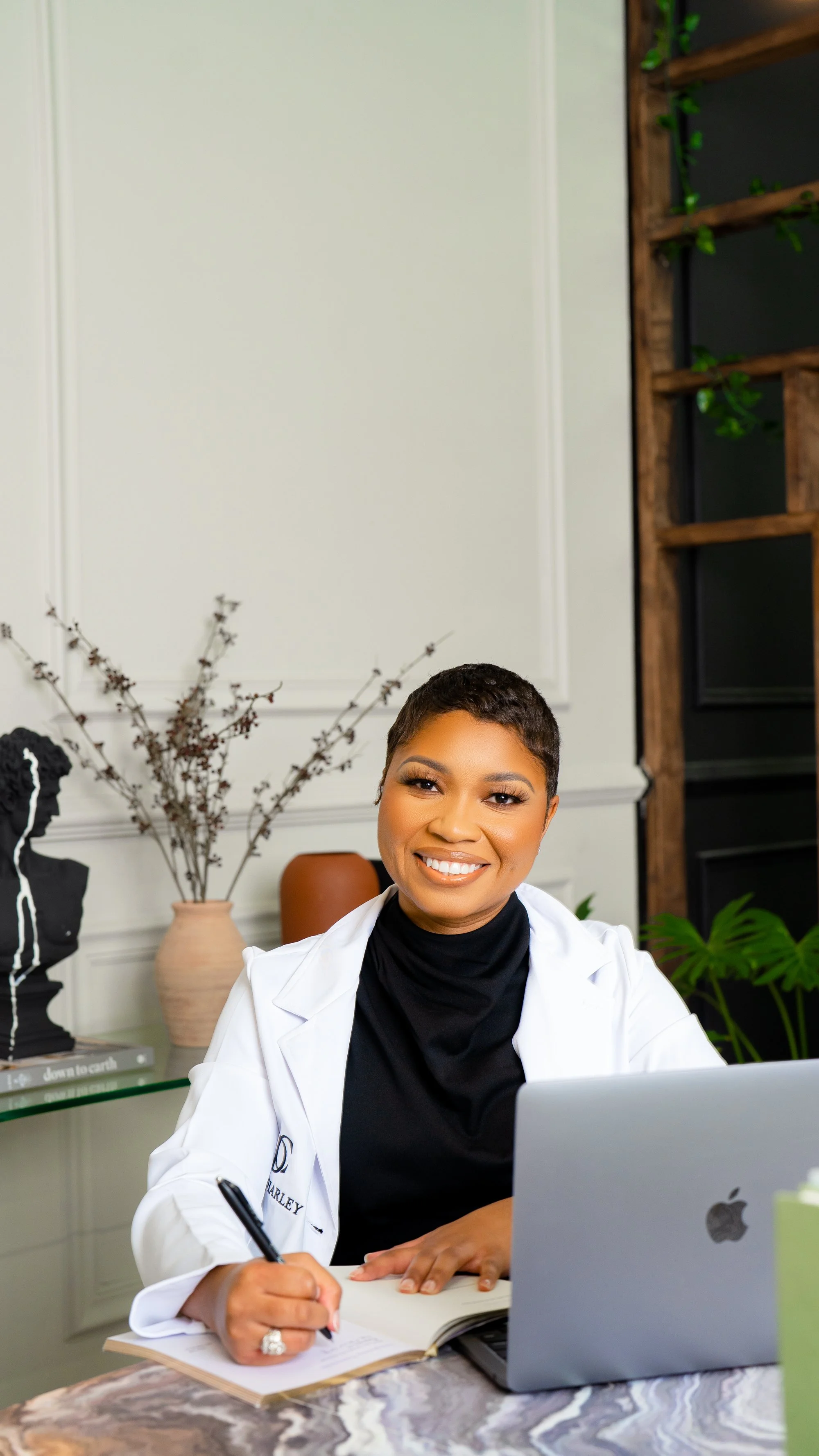 A woman with short hair wearing a black turtleneck and white coat, smiling while writing in a notebook, sitting at a marble table with a laptop in a modern room decorated with plants and art.