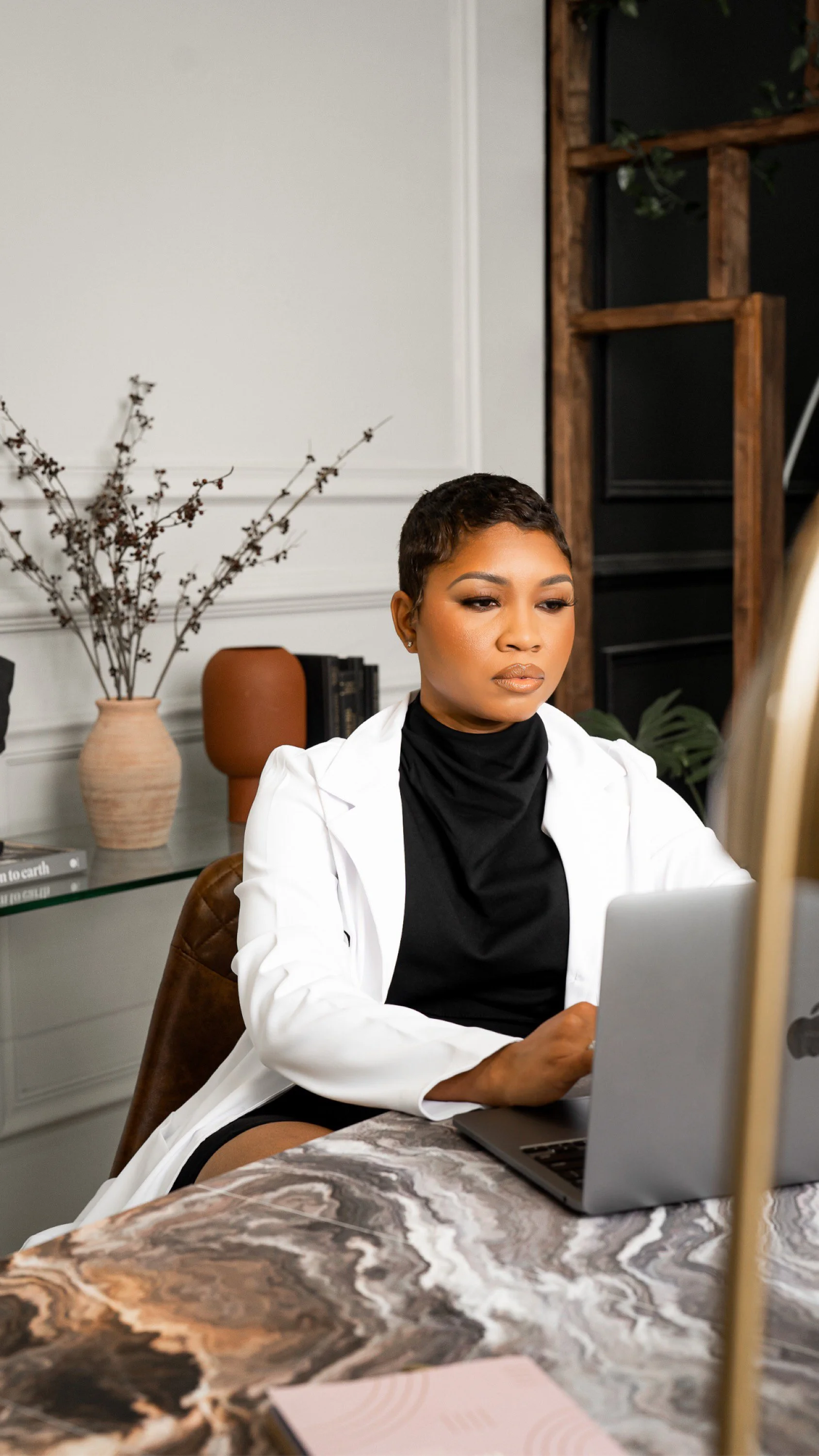 A woman with short dark hair and a black turtleneck sitting at a marble table, looking at a laptop. She is wearing a white blazer. The background includes a vase with dried branches, a bookshelf, and a dark wooden wall.