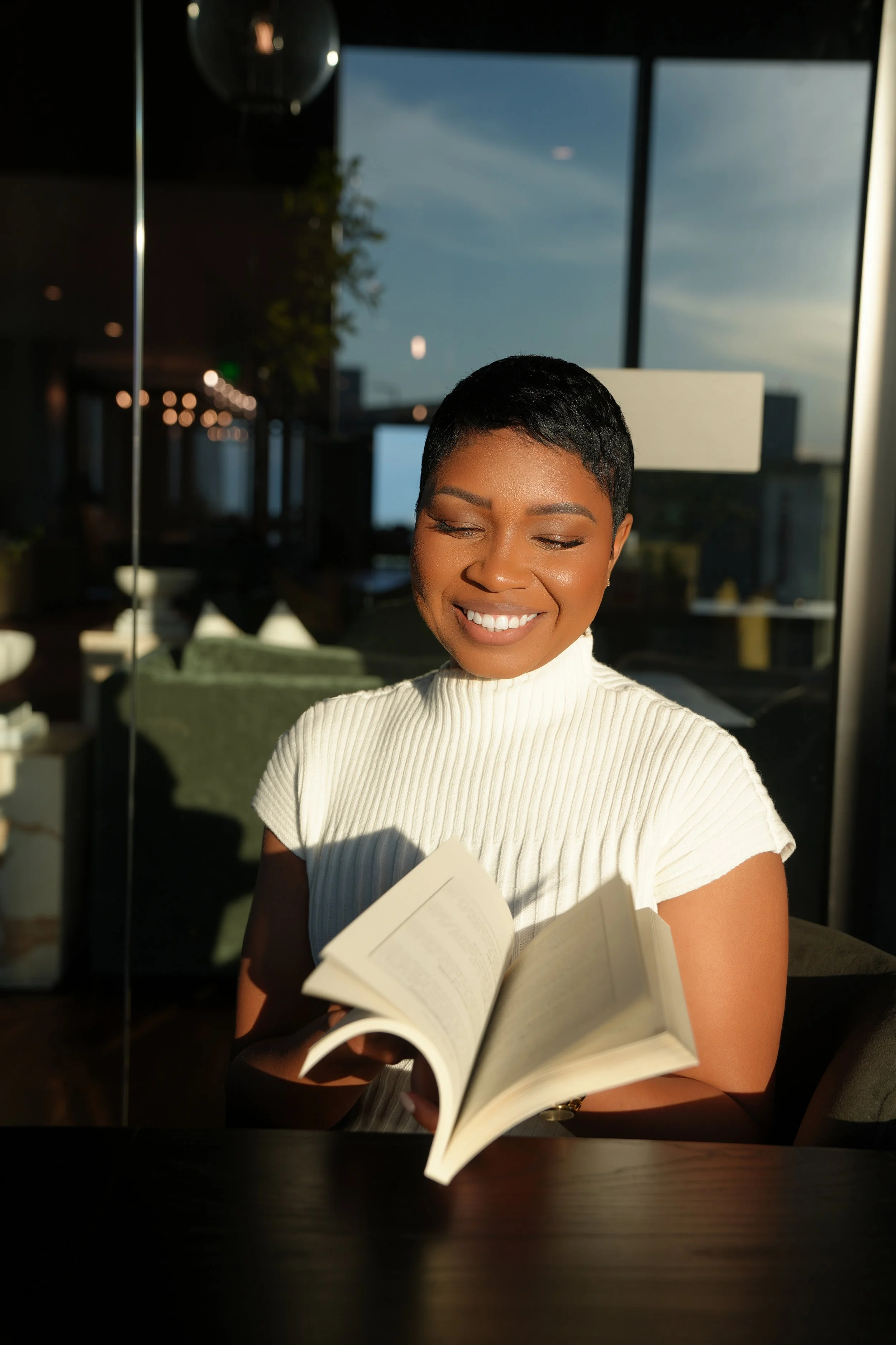 Dr. Charley with the credit card game book in hand and  a bright smile is reading a book in a well-lit indoor space, with large windows showing a blue sky outside.