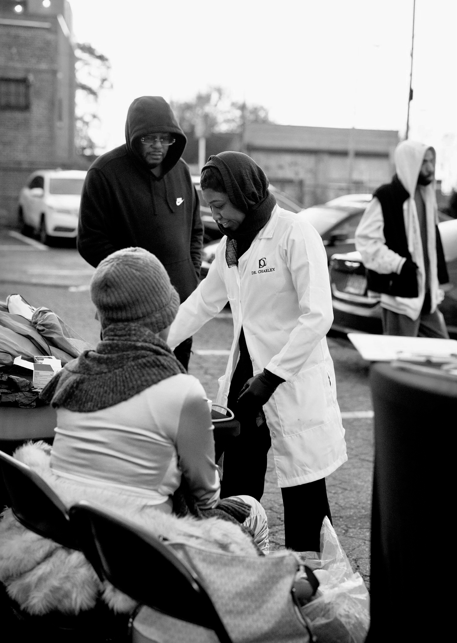 A black and white photo of people at an outdoor event. A woman in a white coat with 'Dr. Charley' embroidered on it is smiling and engaging with seated individuals, while a man wearing a black hoodie with a Nike logo observes. Other people are in the background, including a man in a hoodie and a person with a white hoodie, near parked cars.