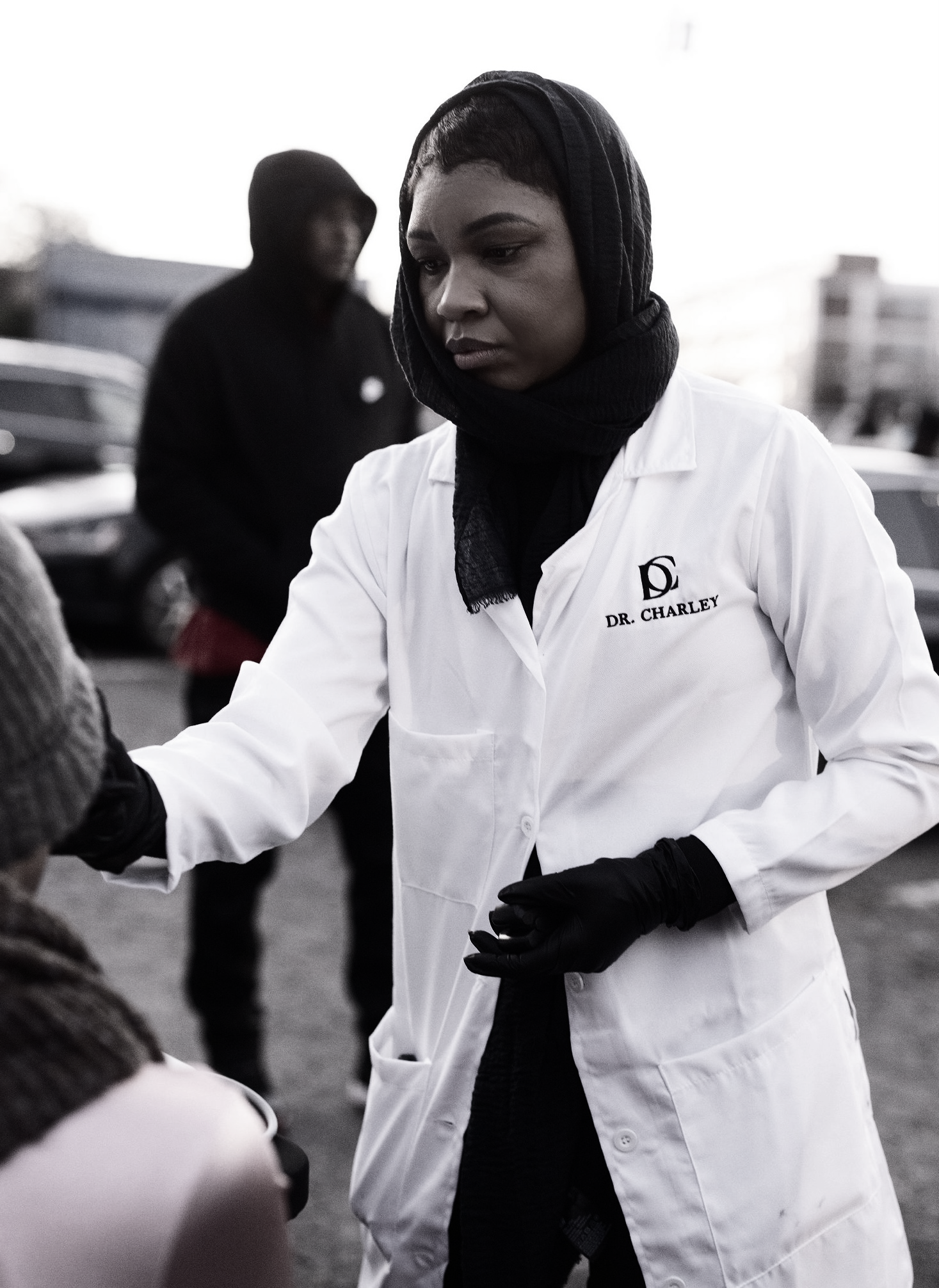 A woman wearing a white coat with 'Dr. Charley' embroidered on it, black gloves, and a head covering, is engaged in a task outdoors. In the background, a man wearing a hoodie is visible, with several blurred vehicles and buildings around them.
