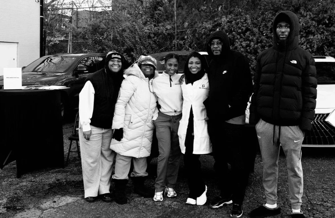 Six people standing outdoors in front of cars, smiling at the camera, dressed in casual and winter clothing.