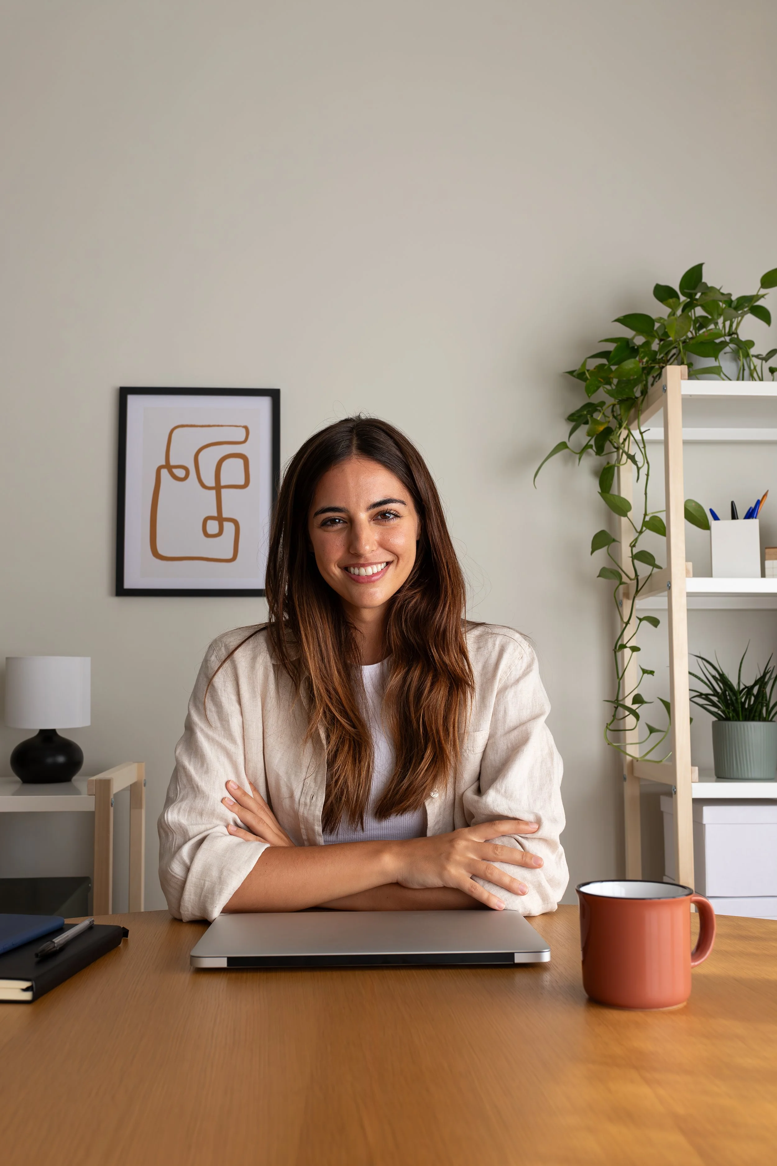 A woman with long brown hair smiling and sitting at a wooden desk in a home office, with a laptop in front of her, a cup on the desk, and shelves with plants and office supplies in the background.