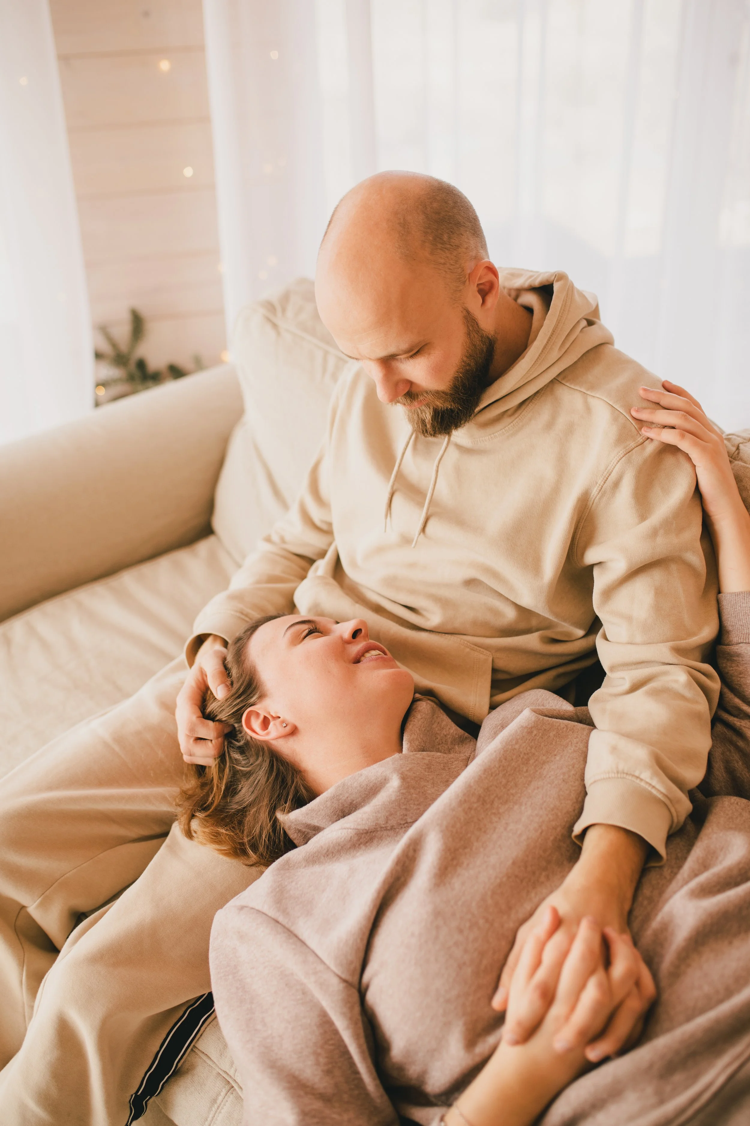 A couple sharing an intimate moment on a couch, with the woman lying down and the man sitting. The woman is smiling and looking up at the man, who is gazing affectionately down at her. The scene appears warm and cozy, with soft natural light coming through a sheer curtain.