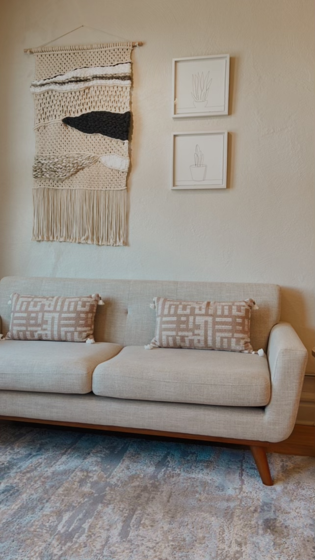 A beige living room with a beige sofa, decorated with two brown patterned throw pillows. Above the sofa, there is a woven wall hanging and two framed line drawings of potted plants. The floor has a light-colored rug with a subtle pattern.
