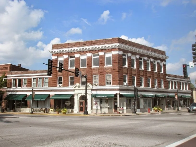 A three-story brick building on a city street corner with storefronts on the ground level and windows on the upper floors, under a partly cloudy sky.