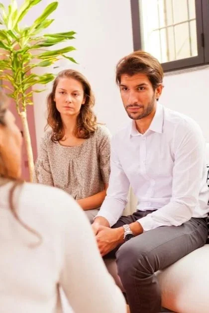 Two people, a man and a woman, sitting attentively during a conversation with a third person, in a room with a large window and a green plant.