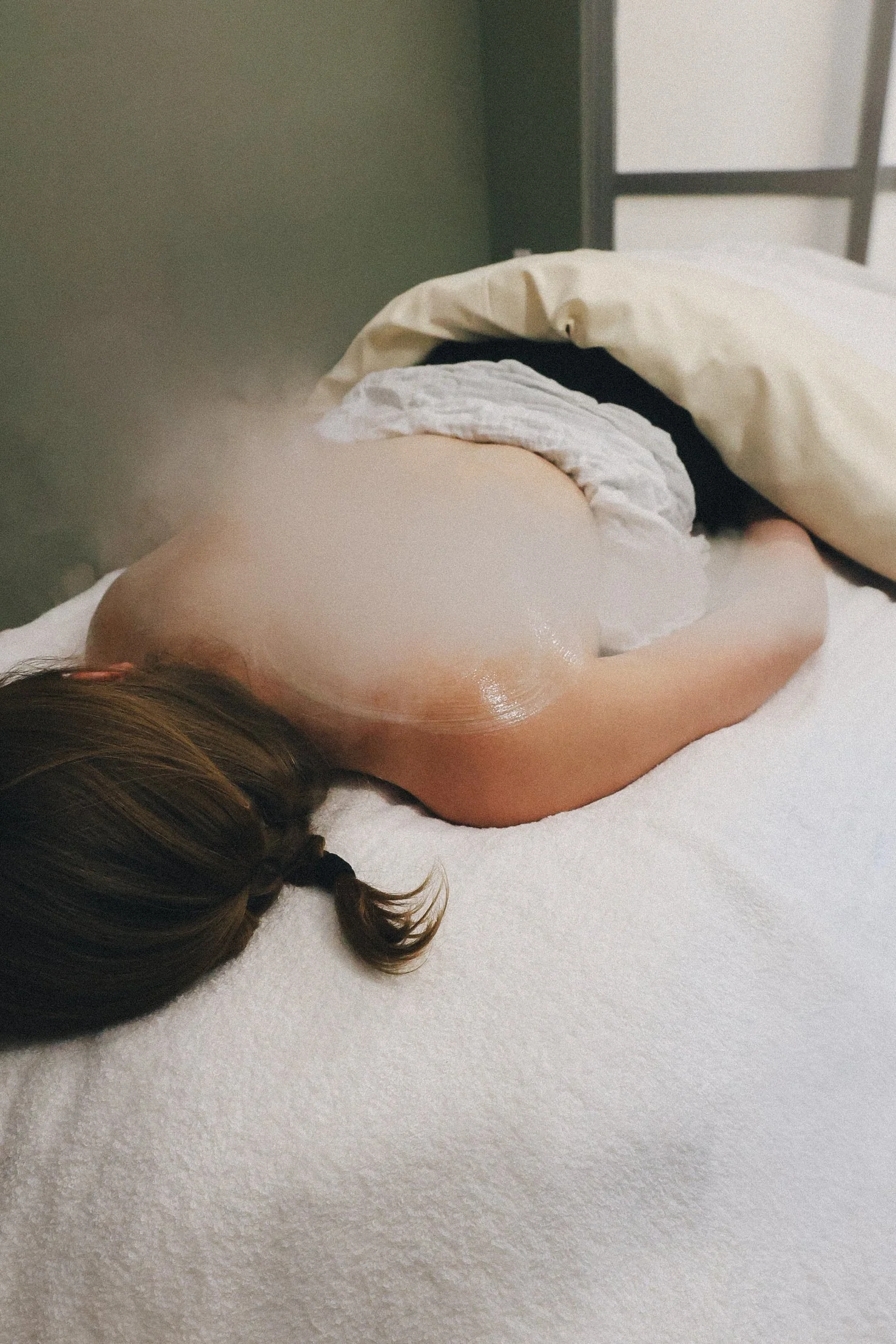A person with brown hair lying on their side on a bed with white sheets, partially covered by a beige blanket, with their back exposed and a shiny adhesive patch on their shoulder.