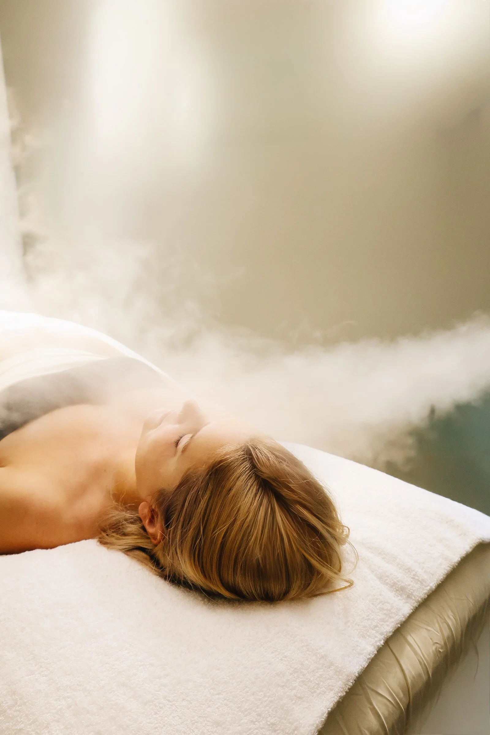 A woman with closed eyes lying on a massage table, receiving a steam therapy treatment in a spa or wellness center.