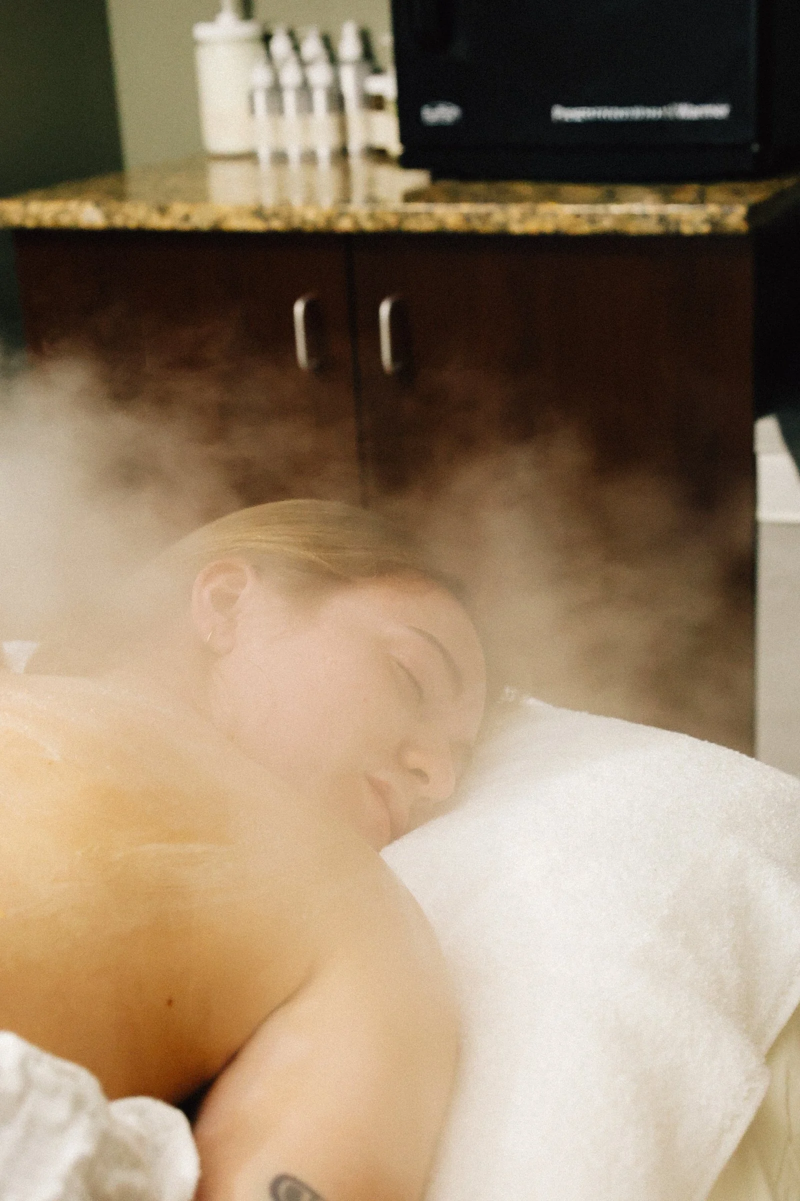 Woman lying on her stomach on a massage table, receiving a steam therapy treatment, with steam around her head.