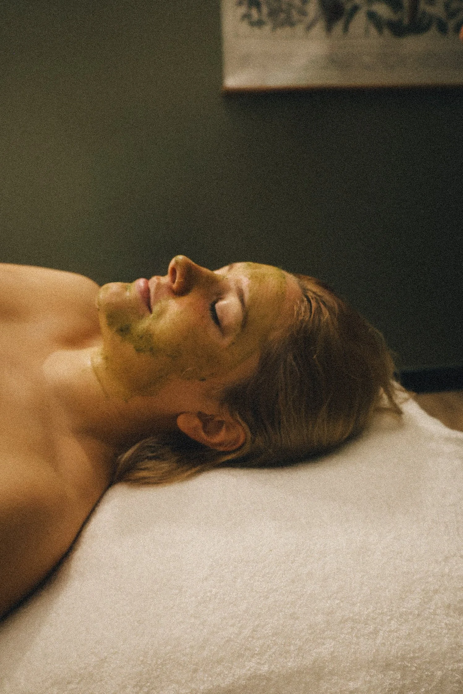 A woman lying on a spa bed with a green facial mask applied to her face, eyes closed, in a relaxing environment.