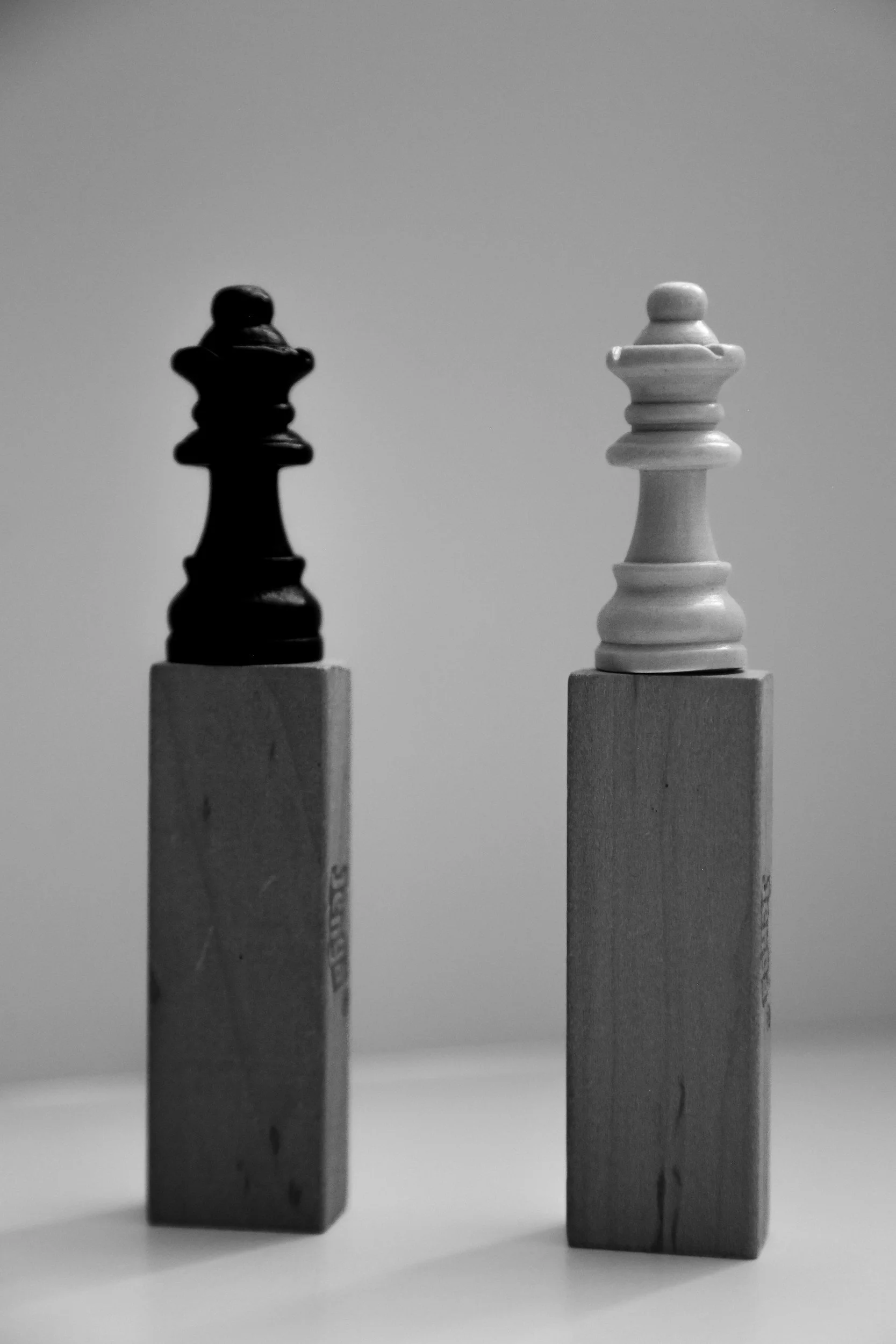 Black and white photograph of a black chess piece and a white chess piece on top of wooden blocks.