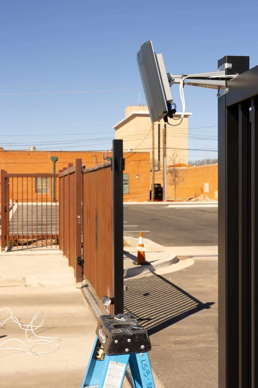Close-up of a security camera or sensor mounted on a black metal gate in an outdoor parking lot, with a brick building and orange traffic cones in the background.