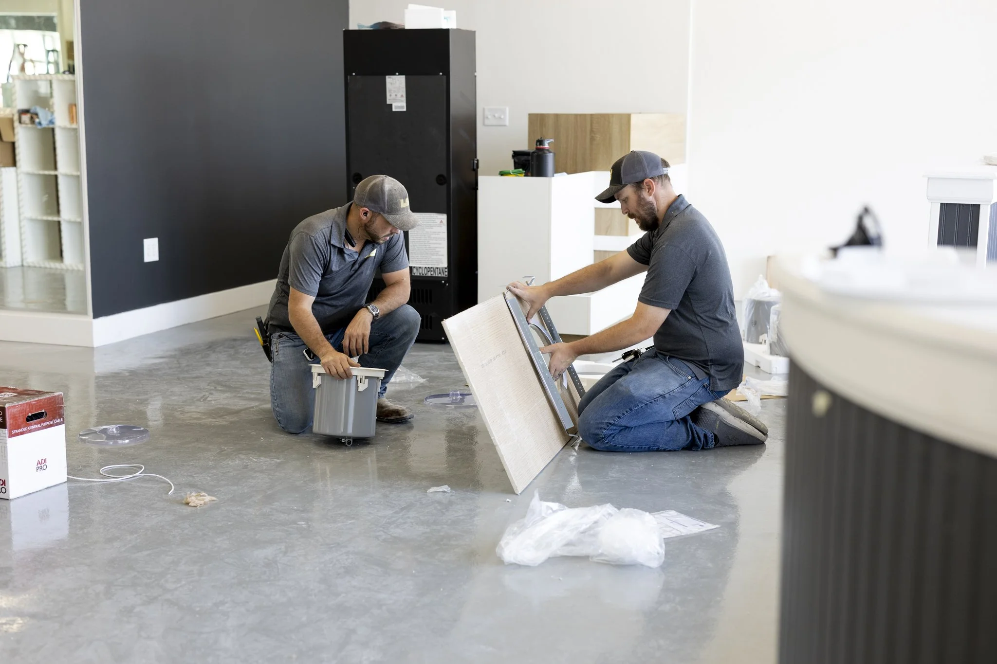 Two men kneeling on a concrete floor installing a ceiling panel in an indoor space.
