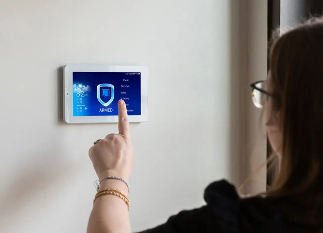 A woman presses a touch screen on a wall-mounted home security system displaying an ARMED status.