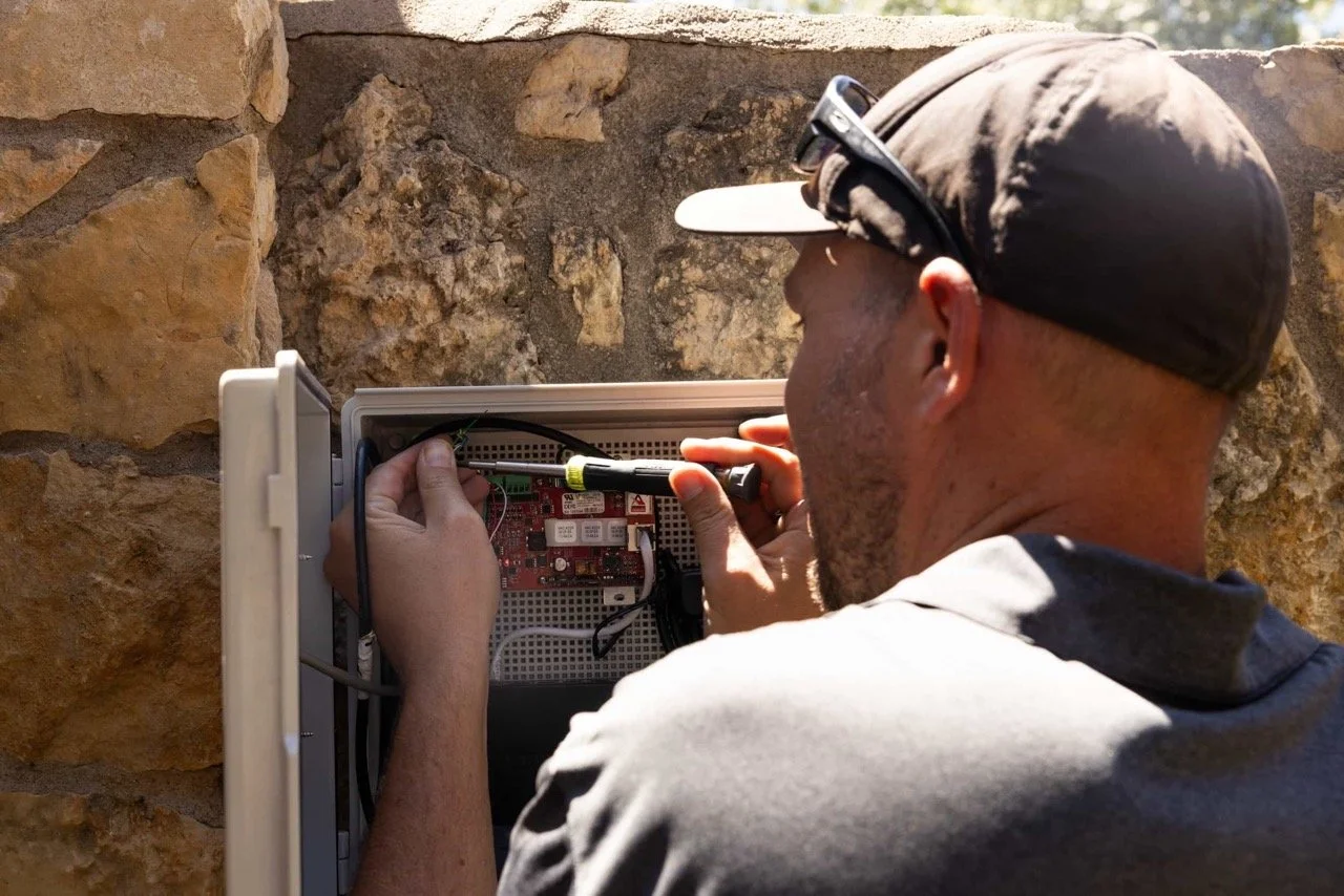 A man in a cap repairs electronics inside an outdoor control box mounted on a stone wall.