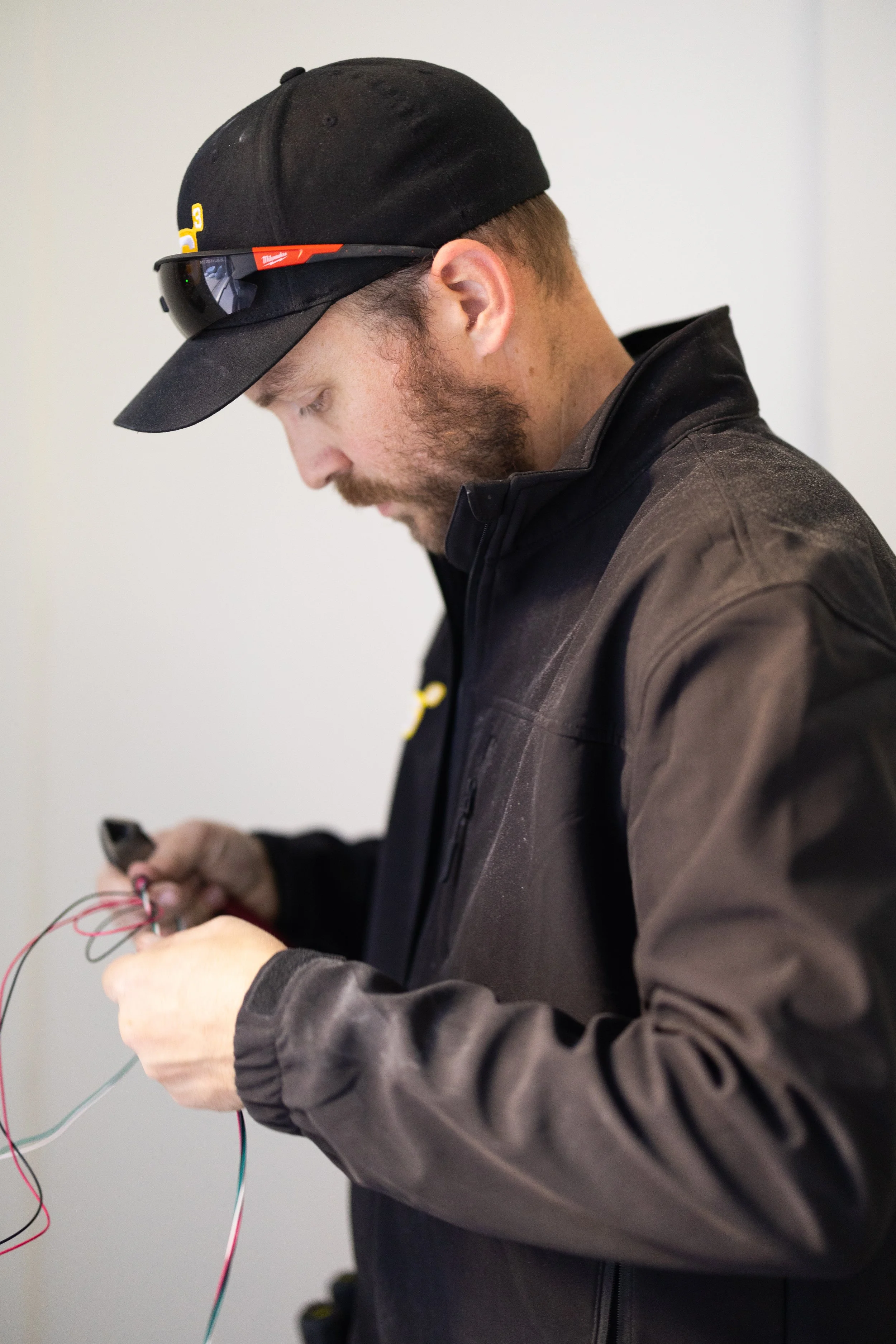A man wearing a black cap and sunglasses on the bill working with wires in his hands, dressed in a black jacket, with a serious focus on his task.