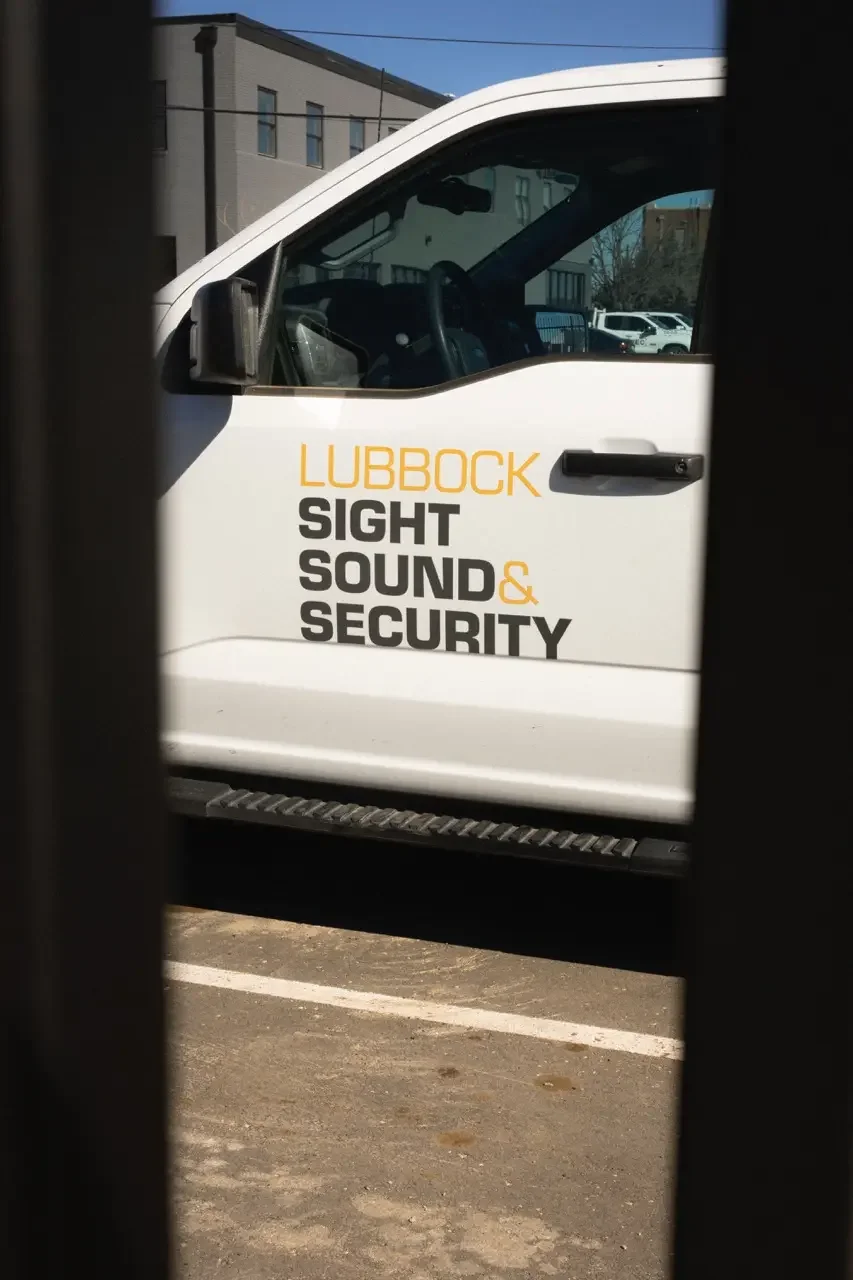White truck with Lubbock Sight Sound & Security logo, partially visible through a dark vertical barrier in a parking lot.