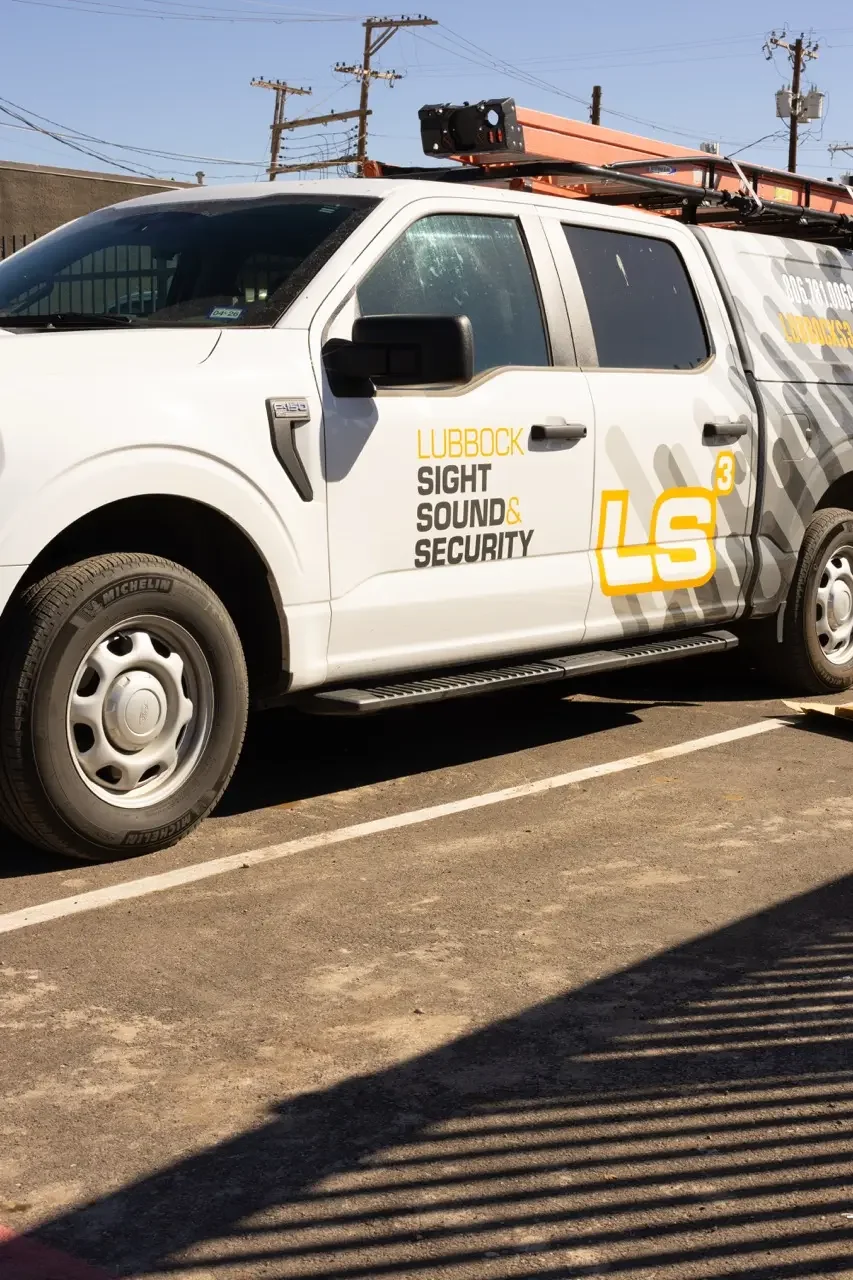 A white work van labeled Lubbock Sight Sound & Security LS³ is parked in a sunlit lot with ladders on its roof.