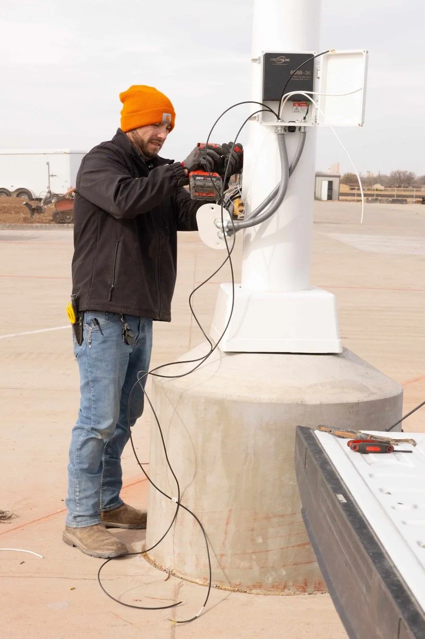 Man in an orange beanie works with wires on a pole-mounted device at an outdoor construction site.