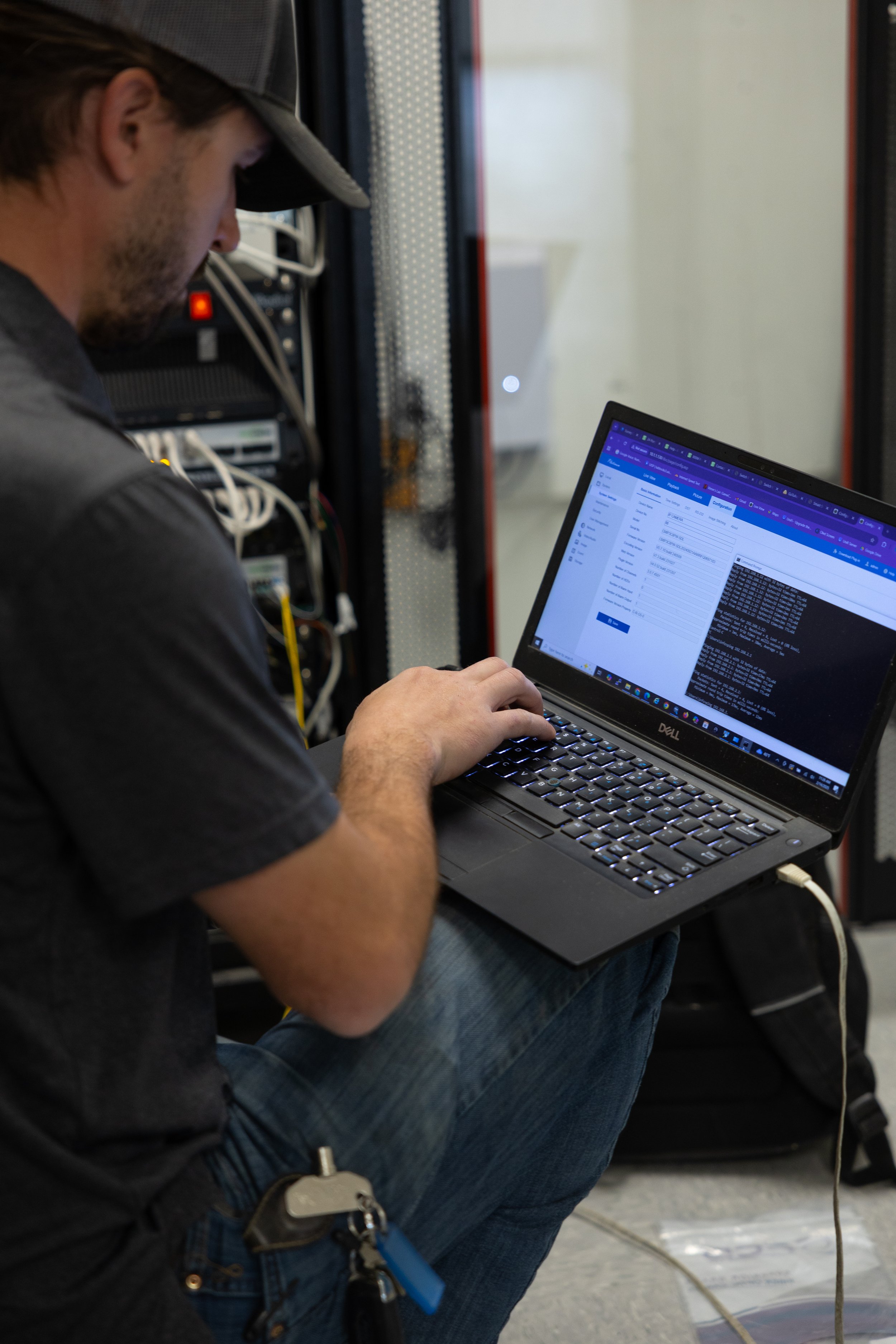 A man wearing a cap and black t-shirt working on a laptop in a server room, with server racks and cables behind him.