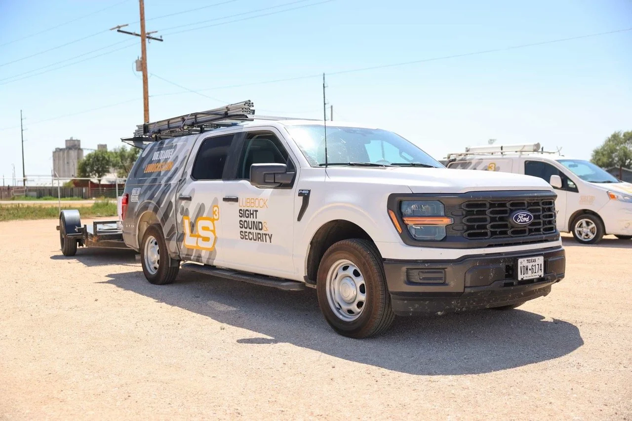 A Ford pickup truck with a light-colored body and black grille, marked with 'Lubbock Sight Sound & Security' and a logo 'LS3'. It has equipment on the roof and is parked on a dirt lot, with utility poles in the background.