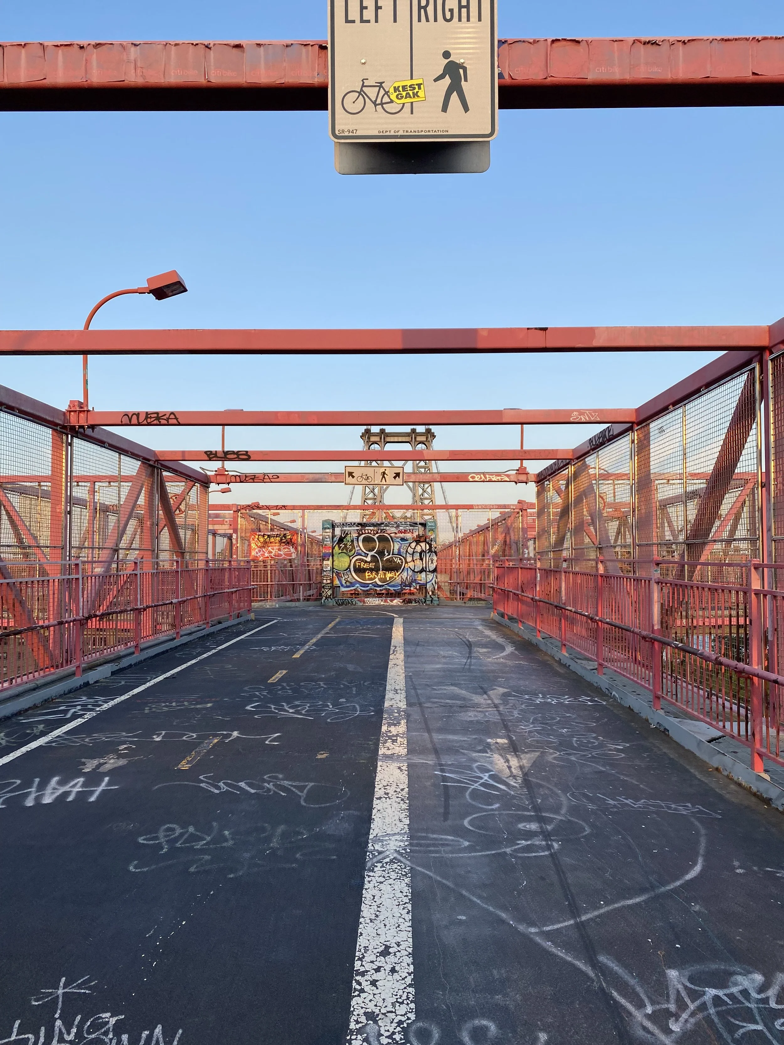 View of a bridge with pink railings and graffiti-covered walls, leading to a structure with graffiti art, against a clear blue sky. Overhead sign indicates sharing the path for bikes and pedestrians with directions for left or right.