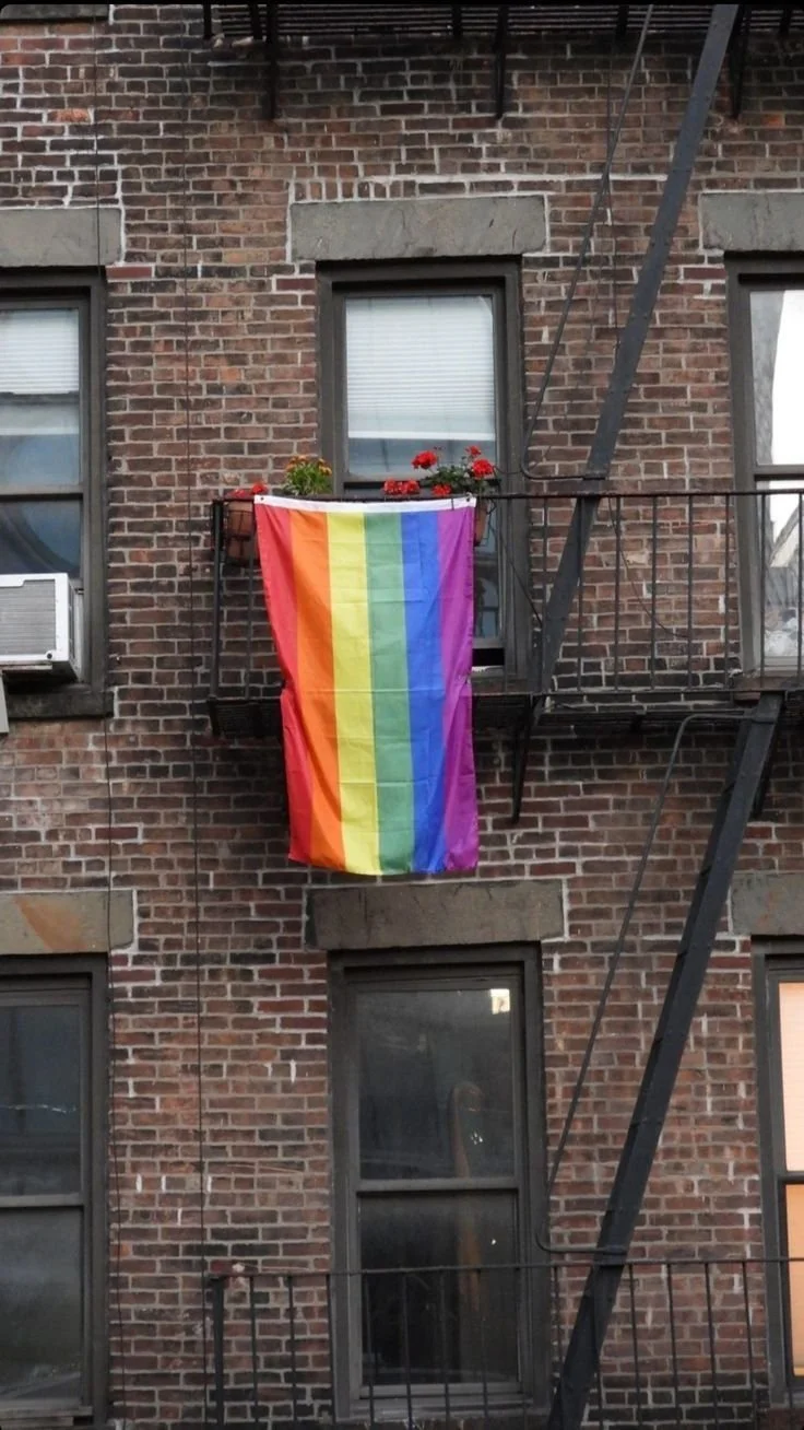 Brick building with four windows, one with an air conditioning unit, and a fire escape. A rainbow flag is hanging from the fire escape on the second floor.