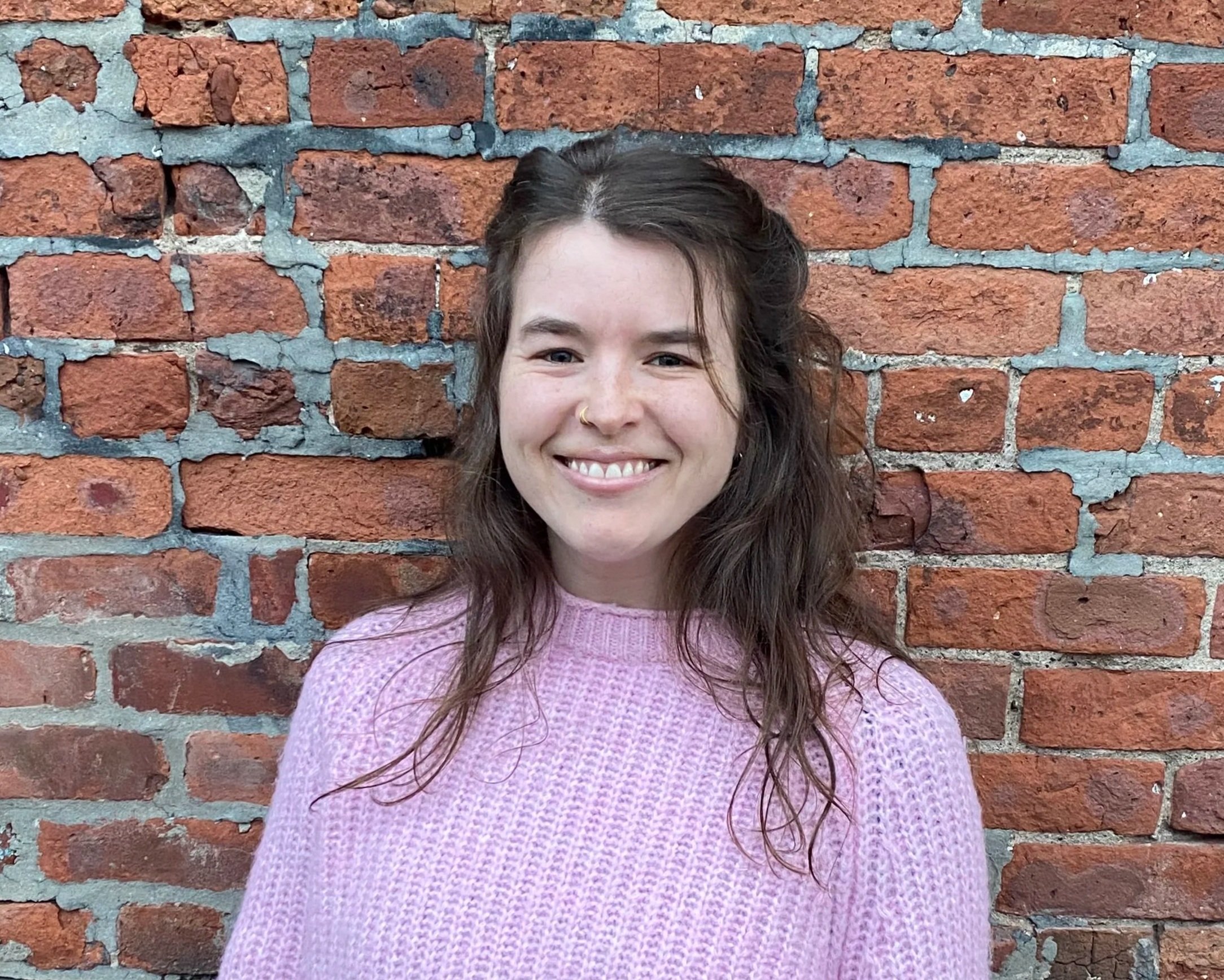A young woman with long, wavy brown hair and a nose ring, smiling, wearing a pink knitted sweater, standing against a red brick wall.
