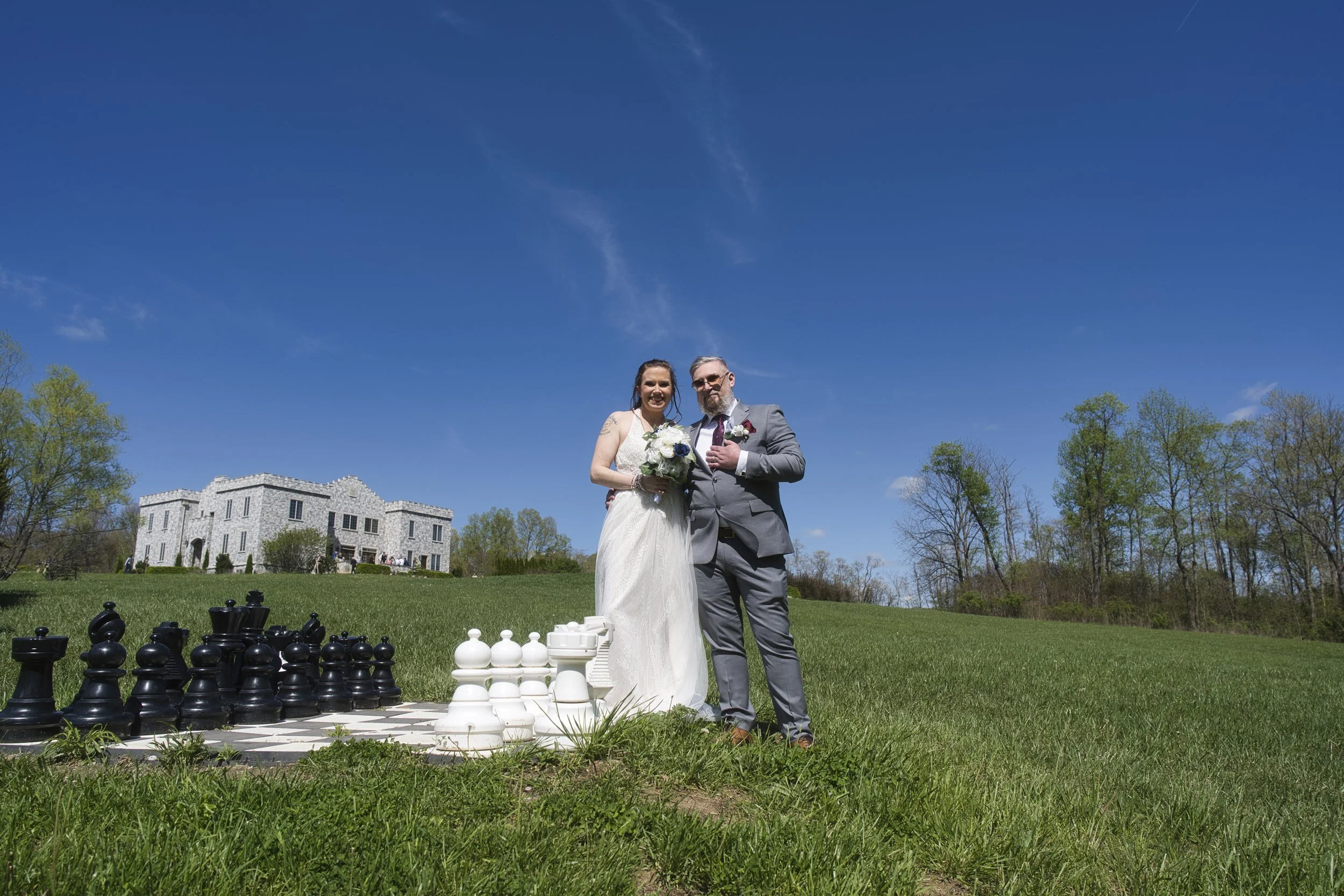 A bride and groom standing outdoors on a green lawn beneath a bright blue sky, with a large chess set on the grass front of them, and a stone mansion in the background.