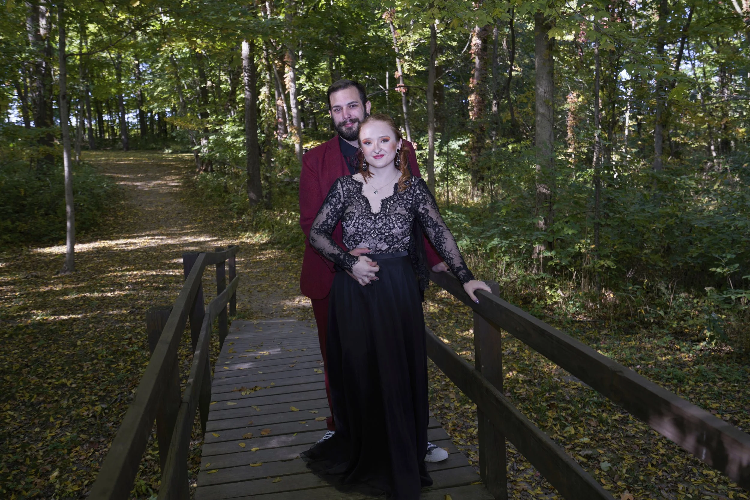 A man and woman standing on a small wooden bridge in a forest with trees and fallen leaves.
