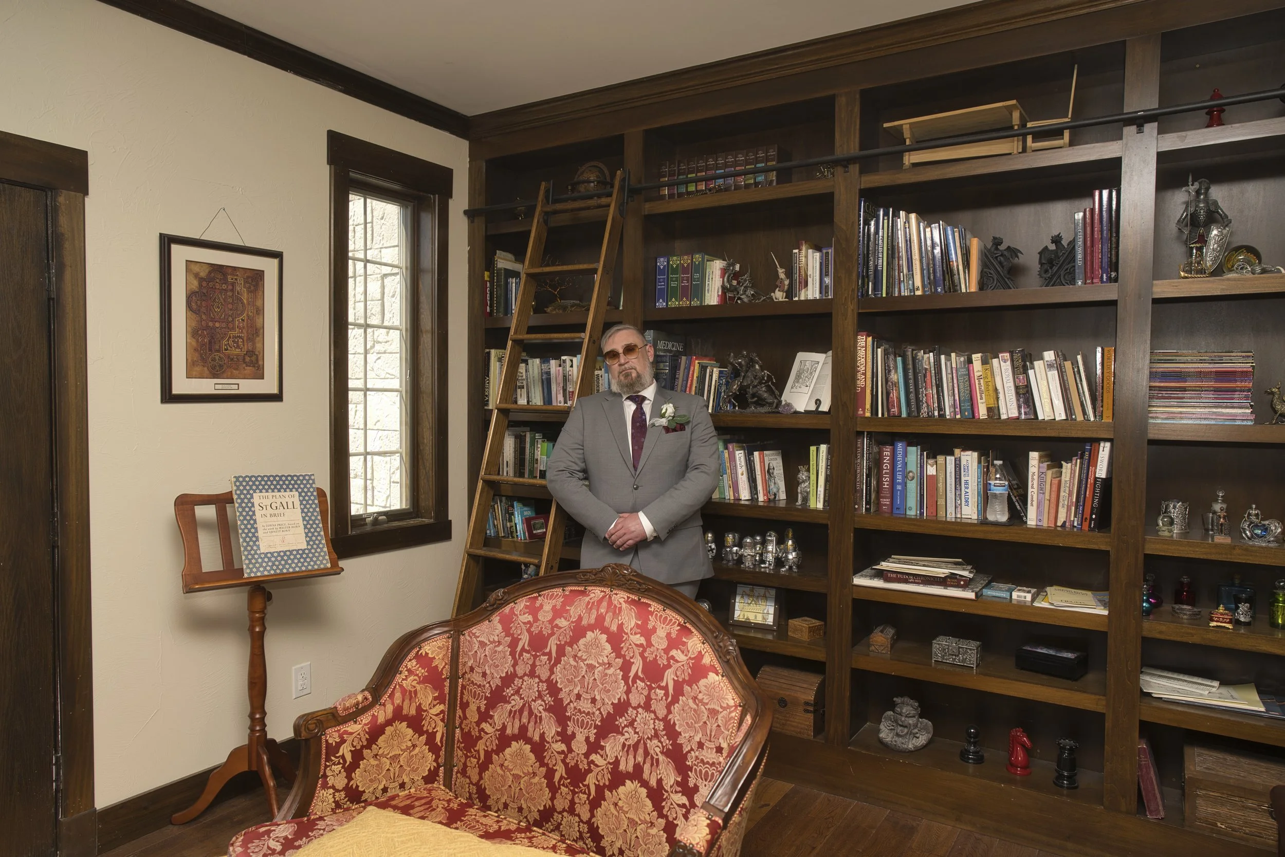 A man in a gray suit with sunglasses and a boutonniere, standing in a room with a large wooden bookshelf, wooden ladder, and floral upholstered armchair.