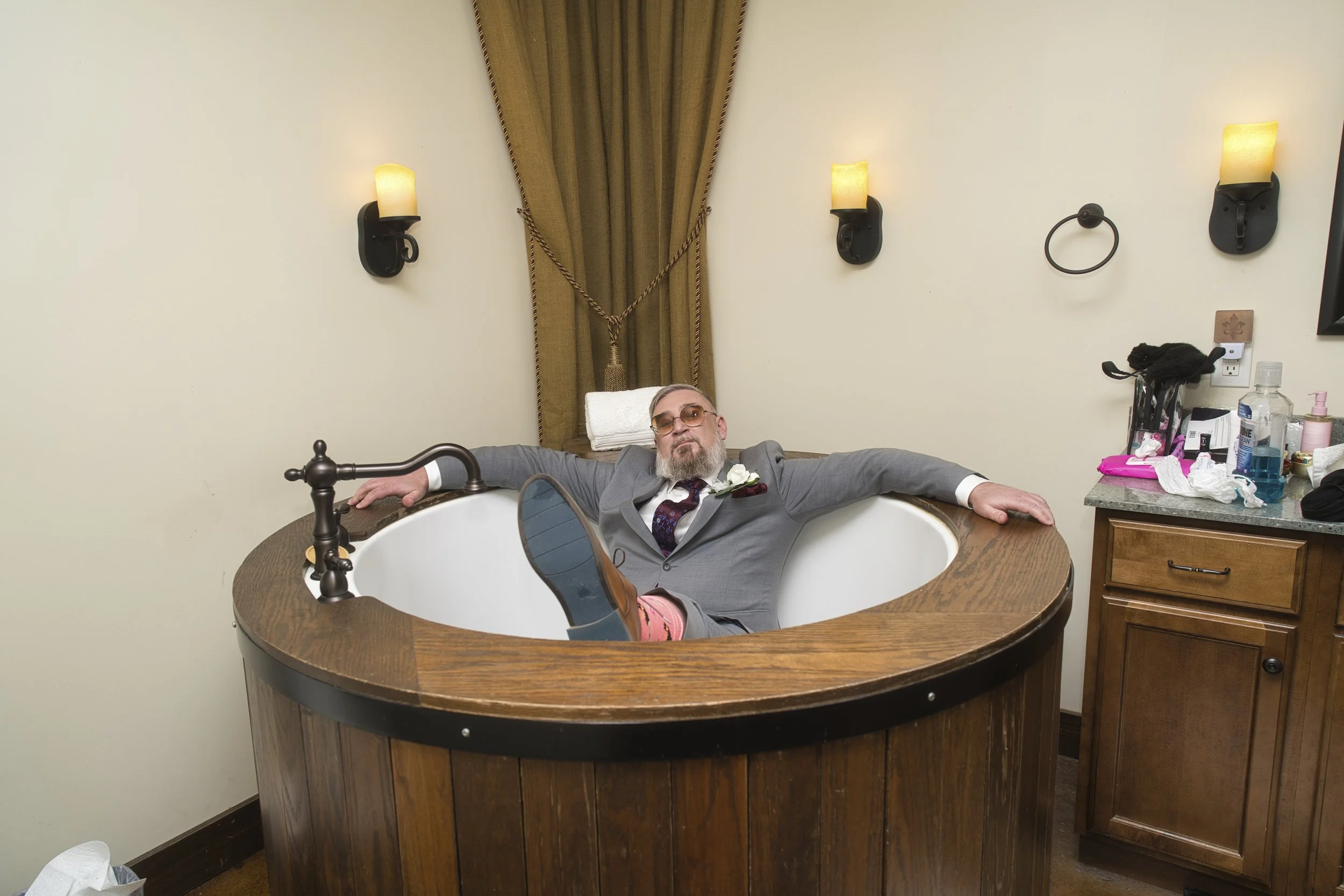 A man in a suit with glasses and a boutonniere is relaxing in a large wooden bathtub in a hotel room, leaning back with arms spread out.