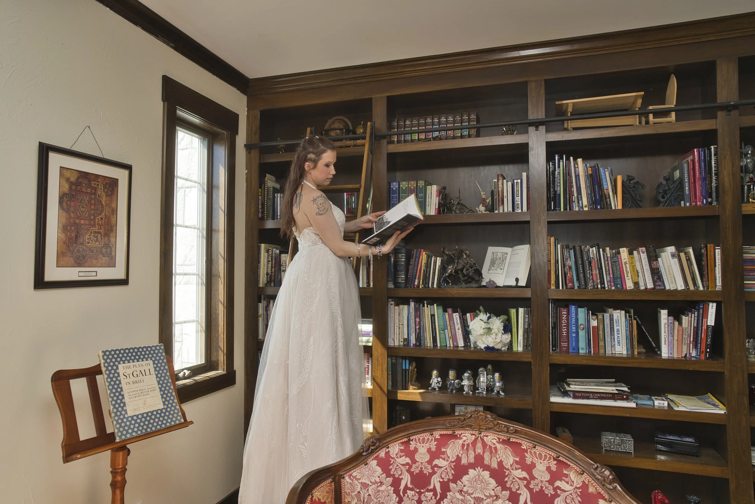 A woman in a white dress reads a book in front of a large wooden bookshelf in a cozy room with a window.