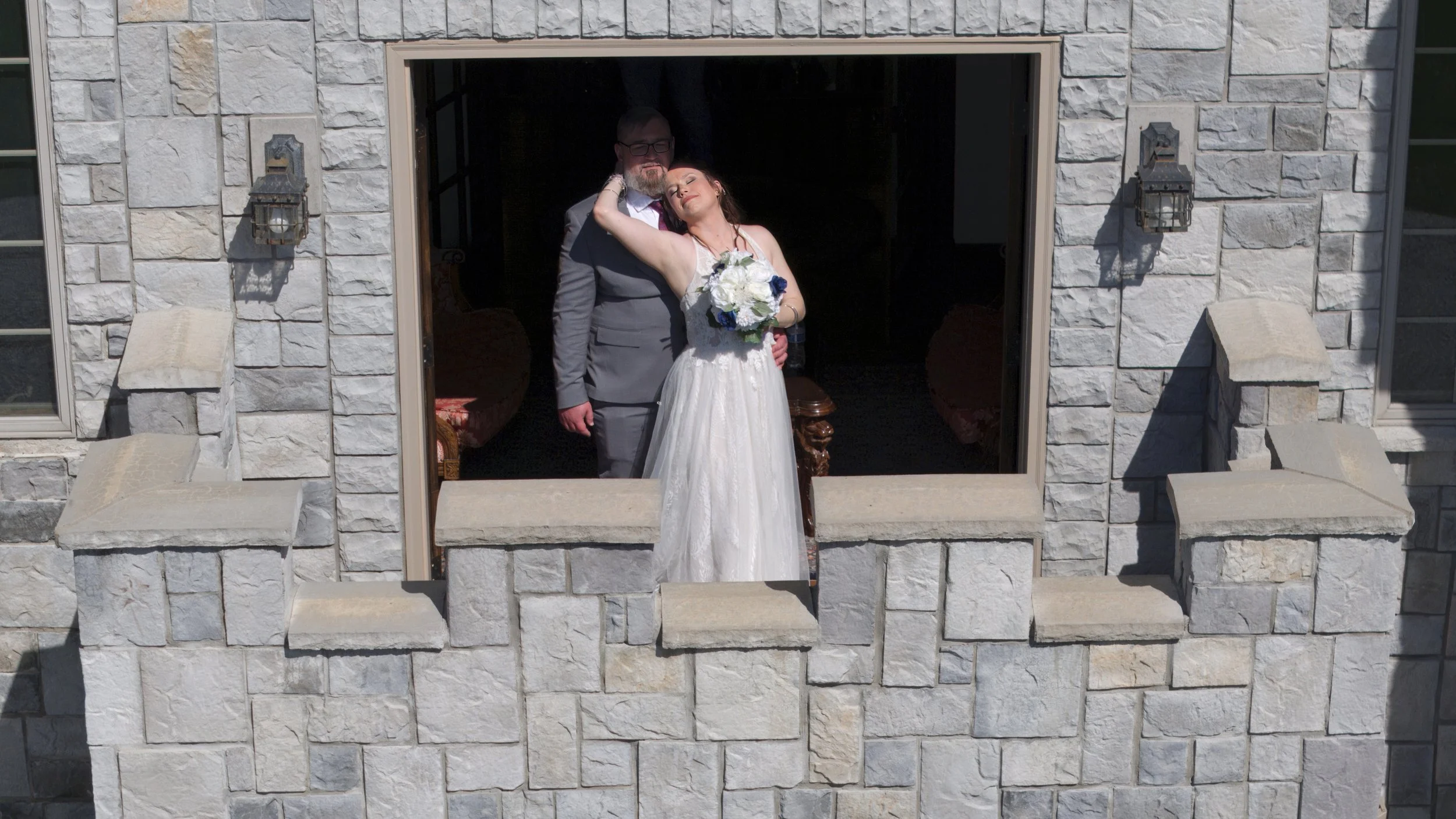 A bride and groom standing on the window ledge of a stone building, during a wedding photo. The bride is wearing a white wedding dress and holding a bouquet, and the groom is in a gray suit with glasses. They are both looking relaxed and loving.