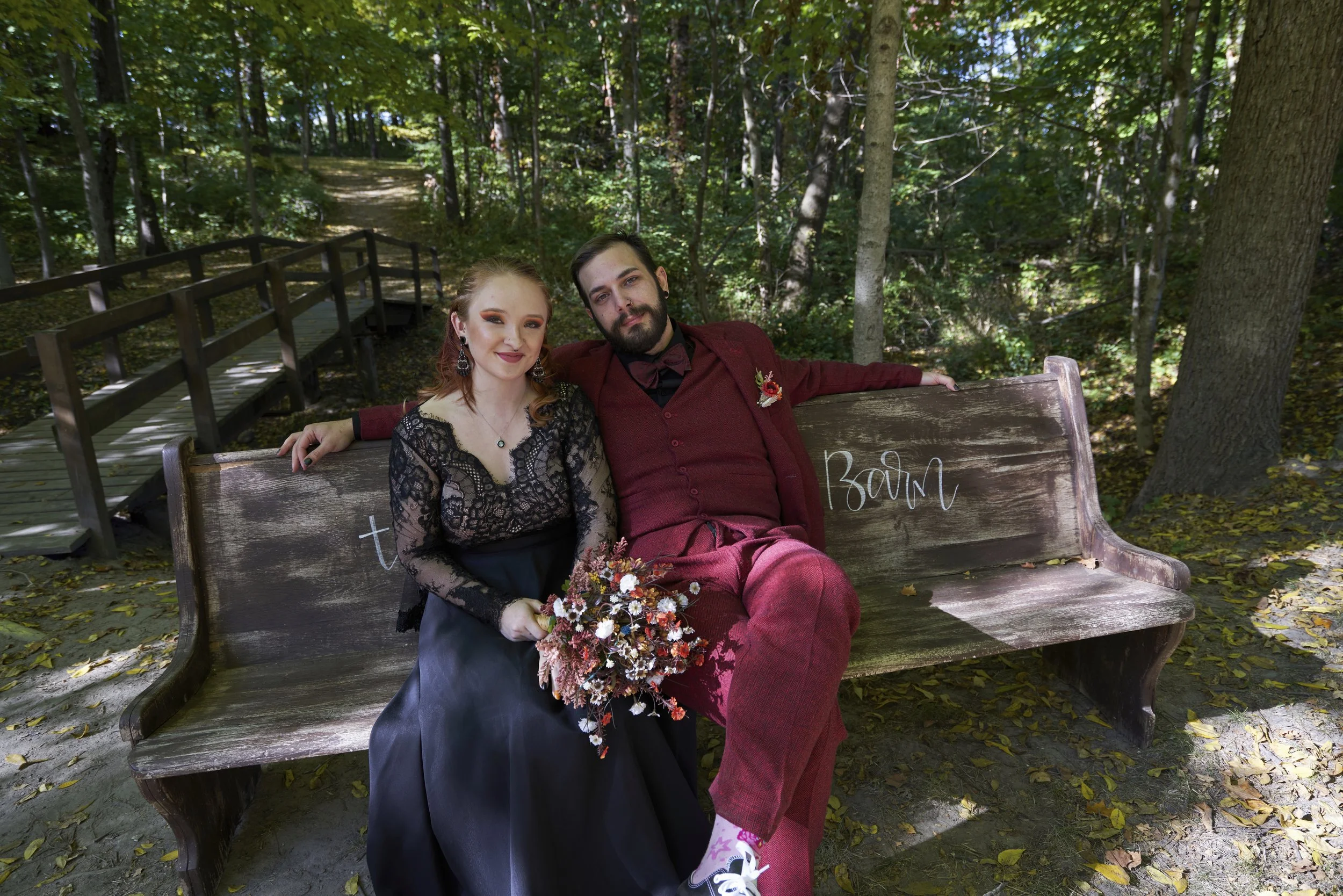 A couple sitting on a wooden bench in a forest. The woman is wearing a black lace dress and holding a bouquet of flowers. The man is dressed in a burgundy suit with a boutonniere. There is a sign on the bench that says "Born". They are sitting closel