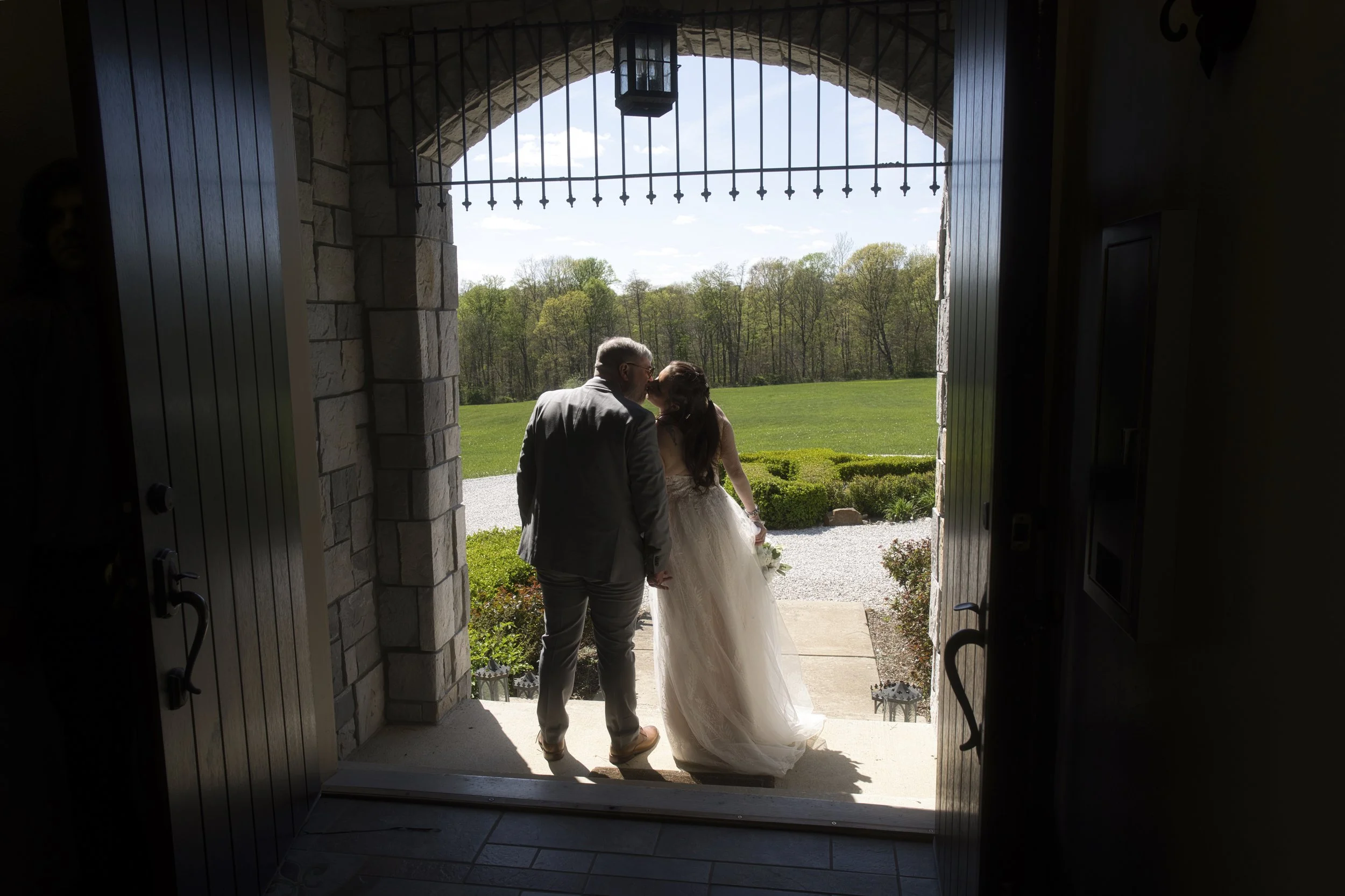 A bride and groom holding hands, standing outside a stone building, sharing a kiss under an archway with a lantern overhead, overlooking a green field and trees.