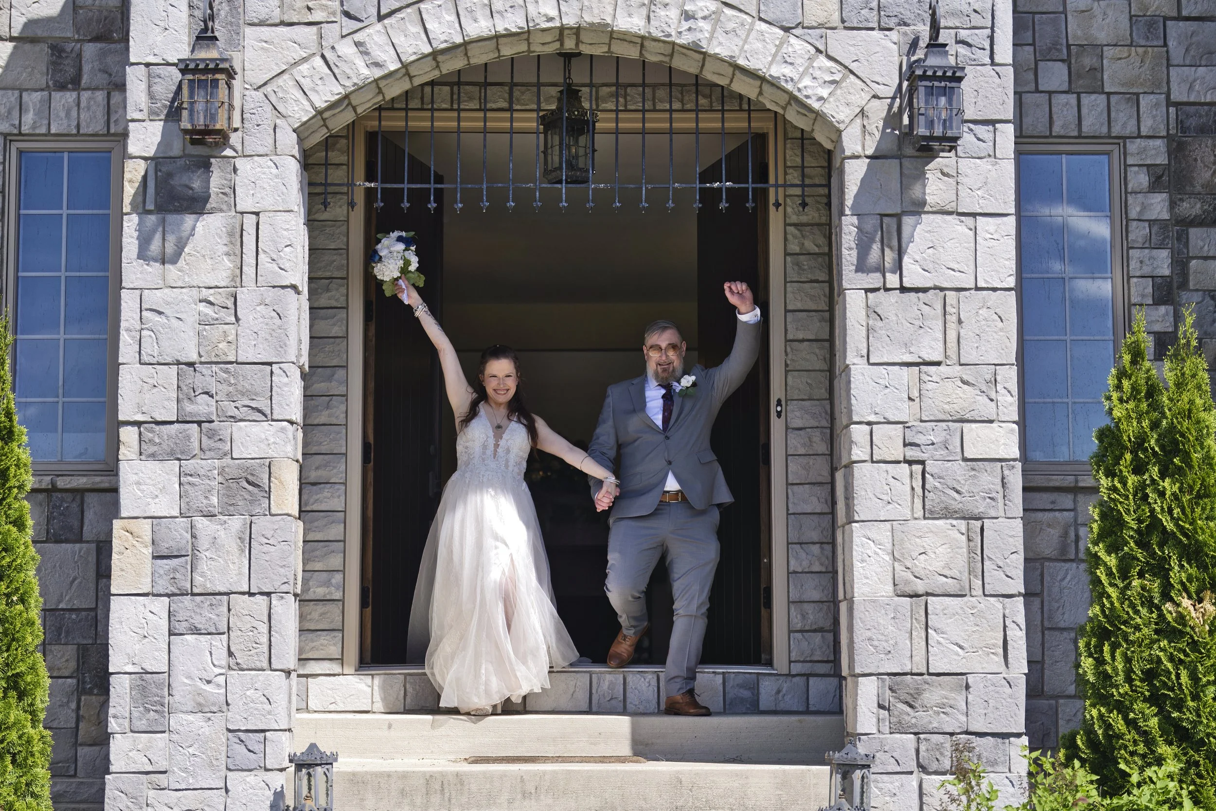 Happy bride and groom celebrating at the entrance of a stone building, holding hands with arms raised in joy.