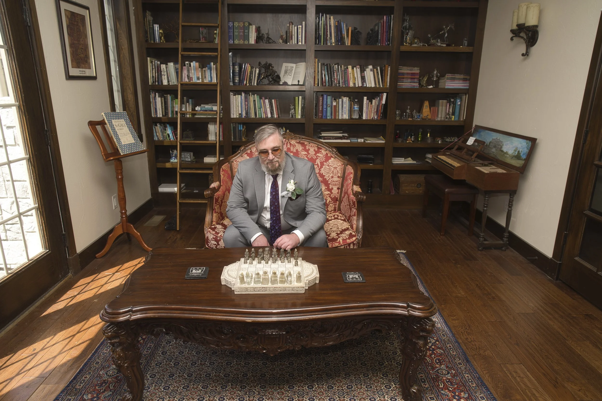 A man in a gray suit sitting on an upholstered chair behind a large wooden table, with a chess set on the table. The room has wooden floors, bookshelves filled with books and decorative items, and sunlight coming through a glass door on the left.