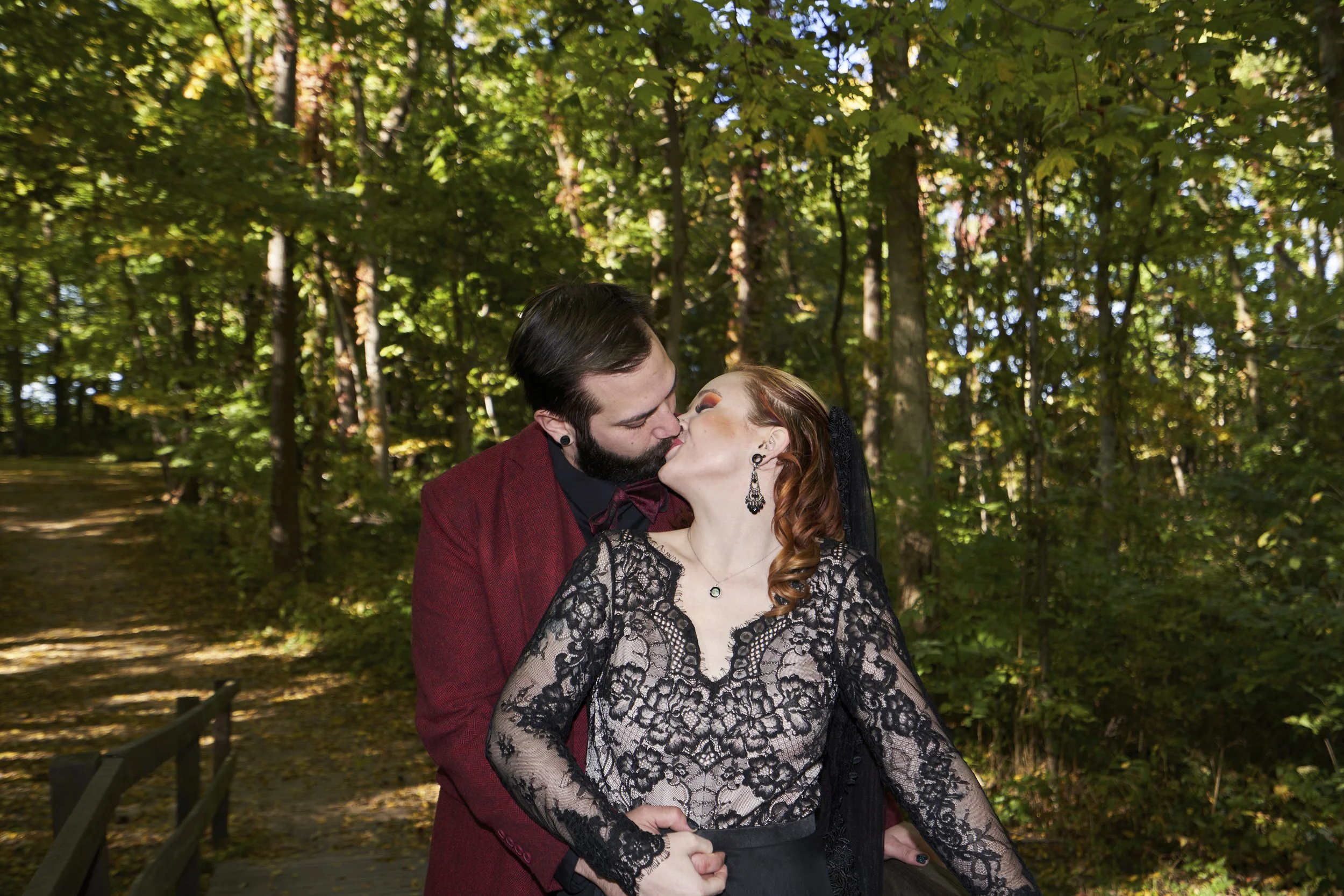 A couple sharing a kiss in a forested area, the woman wearing a black lace dress and the man in a red blazer.
