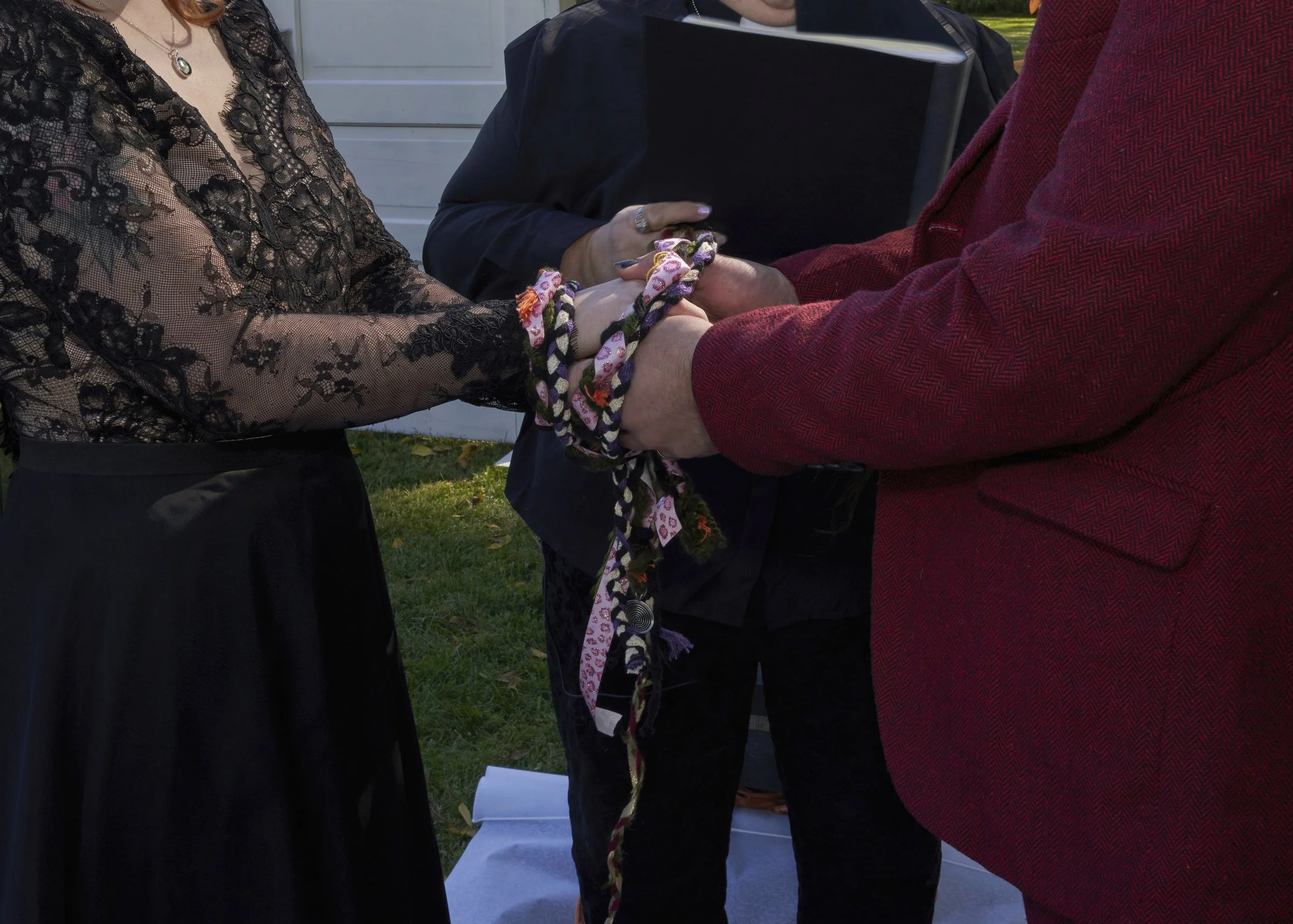 A couple holding hands and tying braided fabric during a wedding or ceremony outdoors, with an officiant in the background.