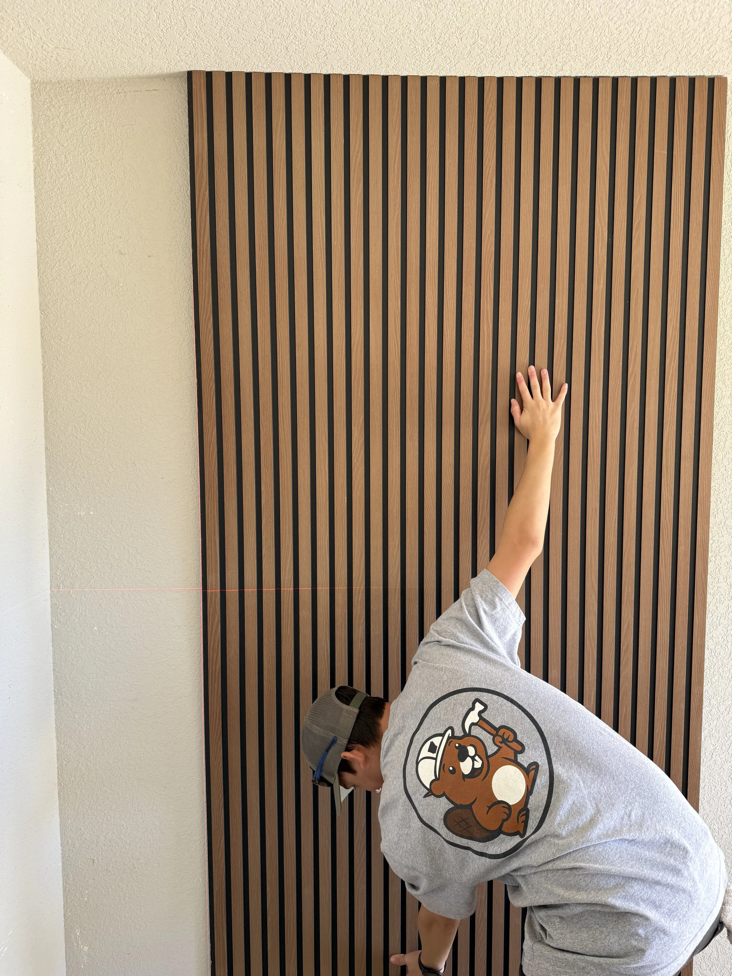 A person wearing a gray T-shirt with a cartoon beaver on the back, a gray cap, and a watch, reaching up with one hand toward a wall with vertical wooden slats.
