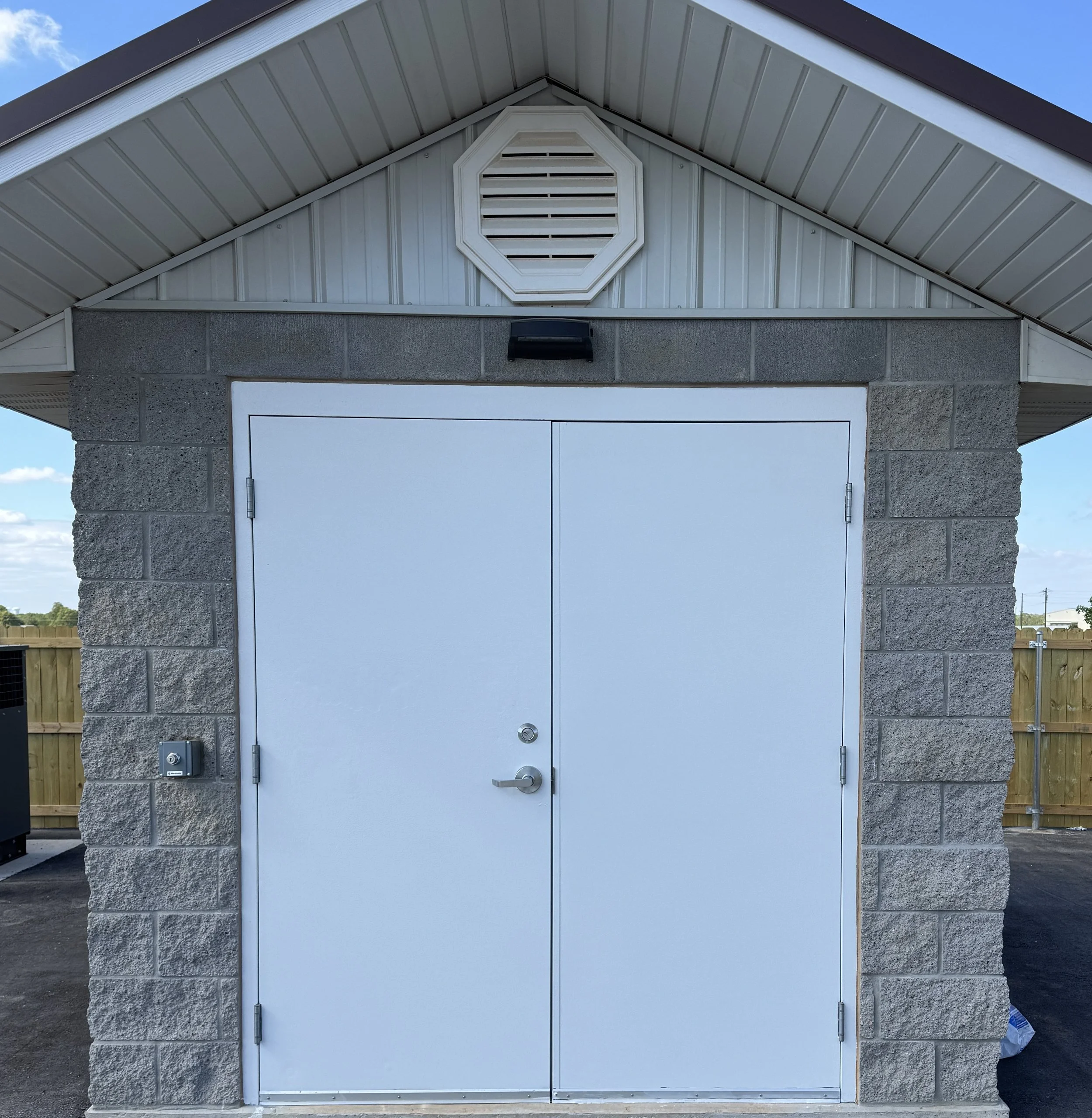 The image shows a corner of a small building with a gable roof, made of grey brick and white siding. There is a white door at the bottom, with a black wall-mounted light above it. An octagonal vent with horizontal slats is located under the peak of the roof. The sky is partly cloudy with a blue background.