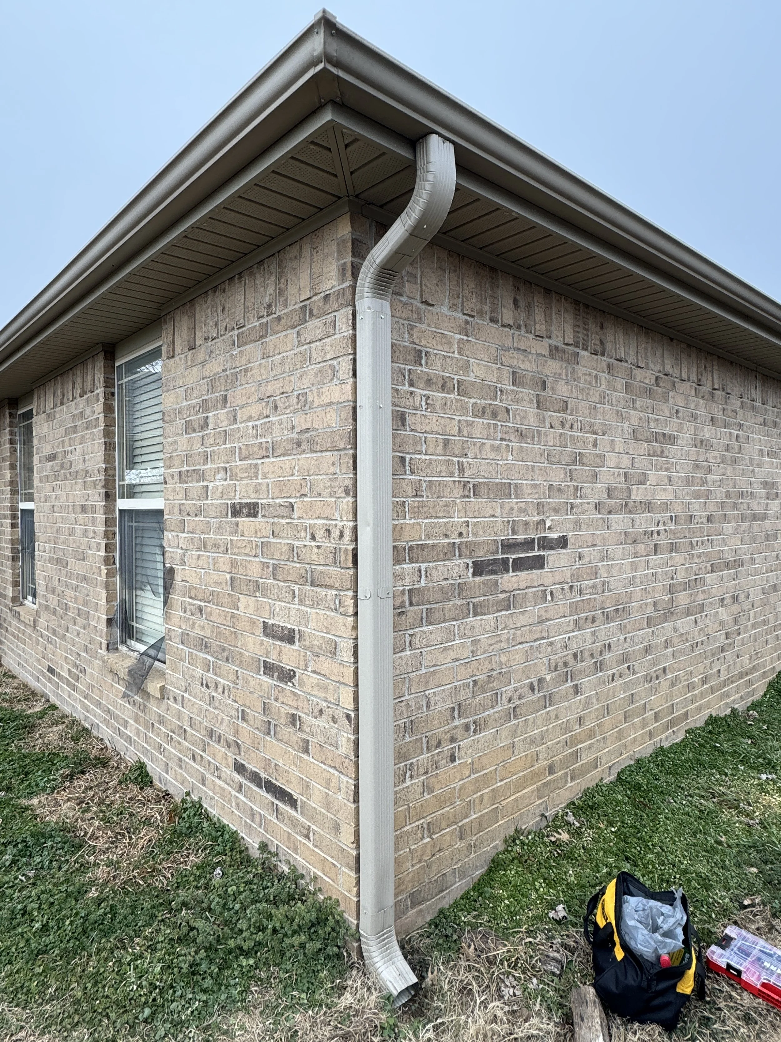 Corner of a brick house with a metal gutter system and downspout, with a grassy area and a yellow and black toolbox on the ground outside.