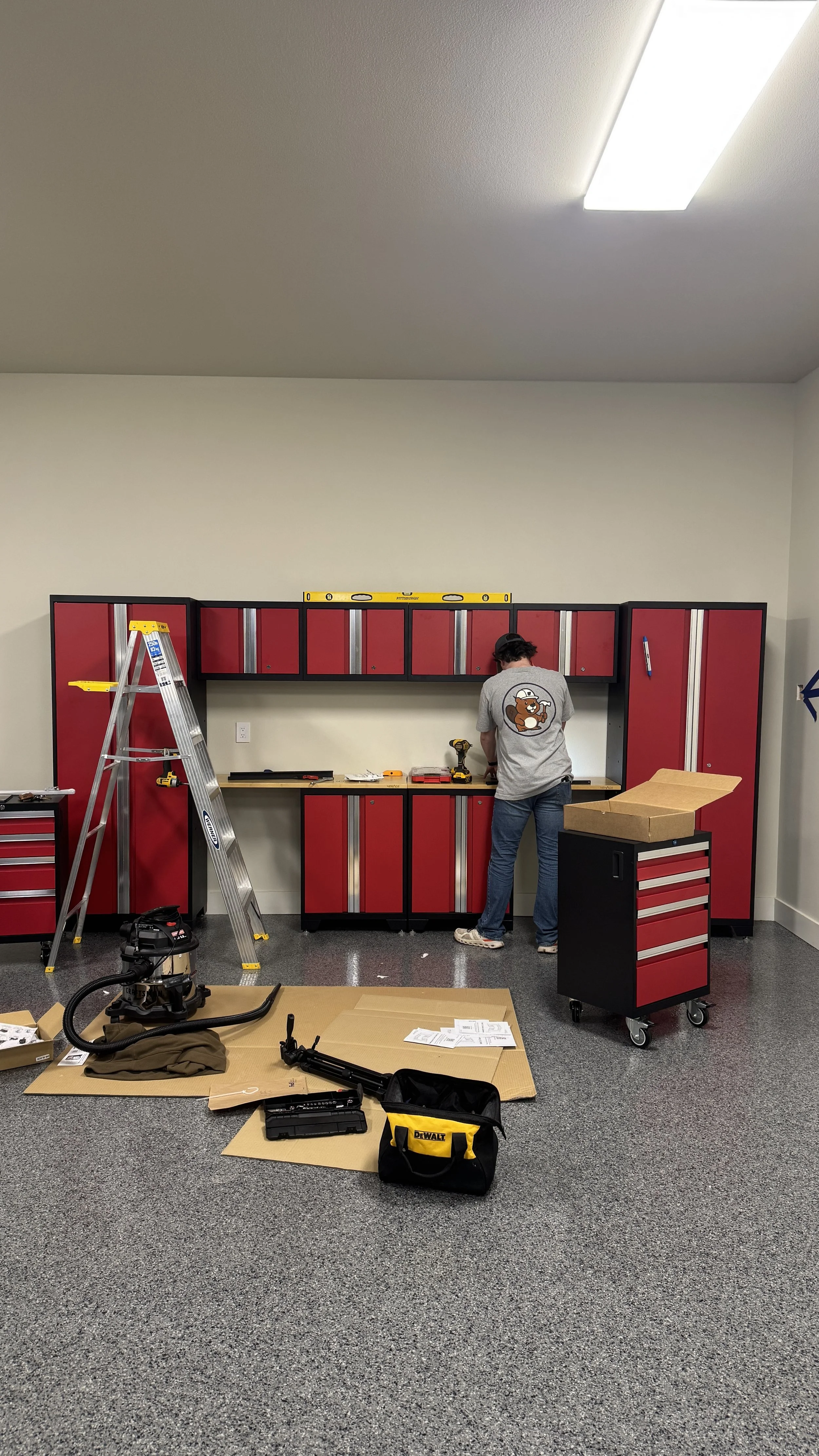 A person working on assembling a red and black garage storage cabinet in a workspace, with a ladder, toolbox, and various tools on a workbench and floor.