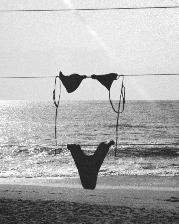 A pair of black bikini tops and bikini bottoms hanging on a clothesline by the beach, with the ocean and waves in the background.