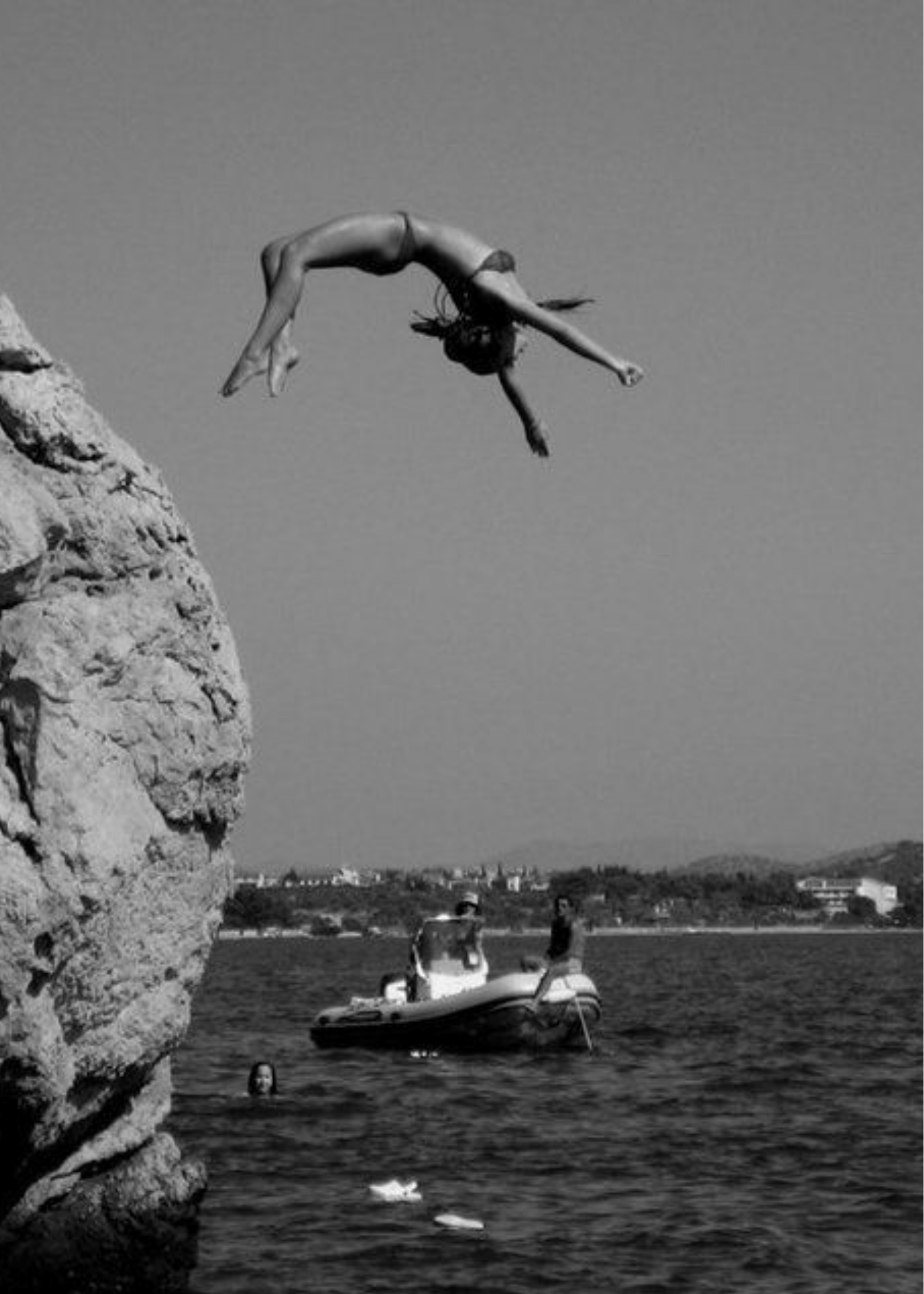 A woman in a swimsuit diving off a rock into the water, with a boat and a person swimming nearby, and a person walking along the shore in the background, in a scenic outdoor setting.