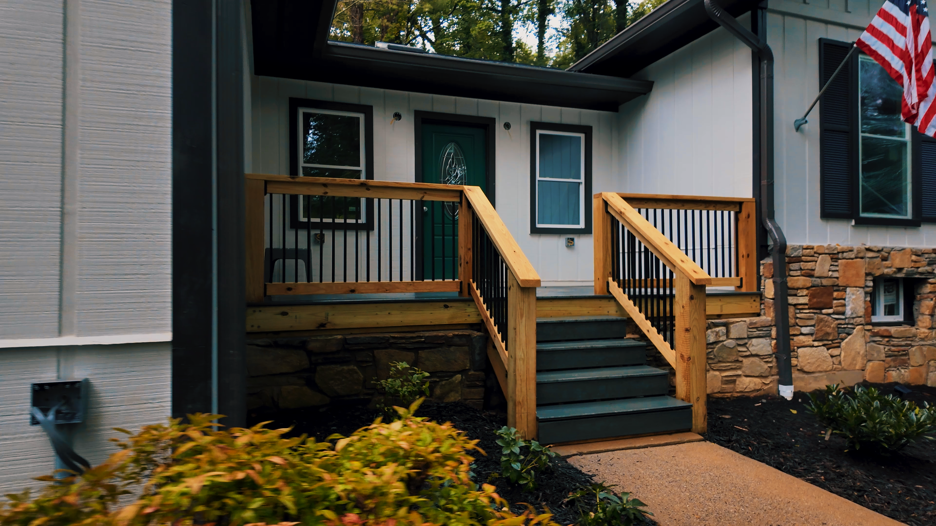 Front porch of a house with a green door and two windows, featuring a wooden staircase with black railings, with an American flag in the yard.