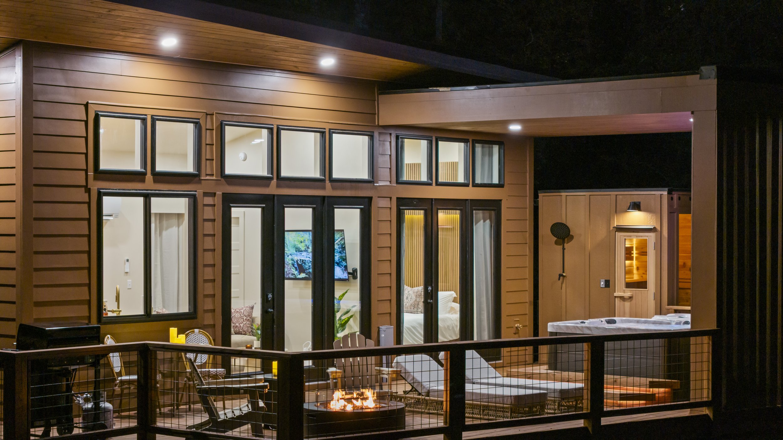 A modern house with large windows and wooden siding, illuminated by outdoor lights at night. There is a balcony with a fire pit, chairs, and a hot tub, with a view into a cozy interior living space.